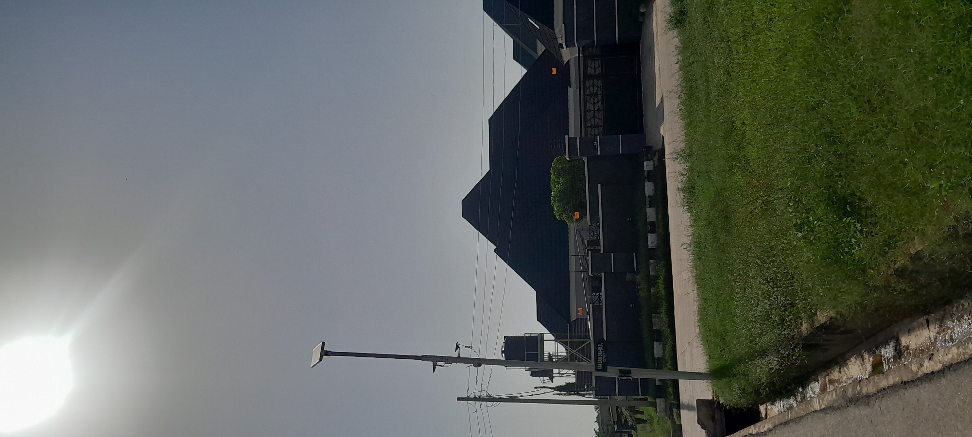 The image captures a sunlit street scene in Jalingo, Nigeria. In the foreground, a paved road runs along the left edge, bordered by a concrete drainage channel and a grassy verge. A tall, grey utility pole with attached equipment, including a solar panel at the top and water tanks supported by a metal structure, stands prominently on the right side of the road. A street sign labeled "YAA ZAKI DAI-HABU STREET" is affixed to this pole.
Behind the utility pole, a modern, dark-roofed residential building with a dark grey perimeter wall and ornate gate is visible. The building features several peaked roof sections. A dense green hedge is situated in front of the wall. The sky above is bright with a strong sun, casting a glare and creating a slightly hazy atmosphere. The overall environment suggests a developing urban or suburban area.
