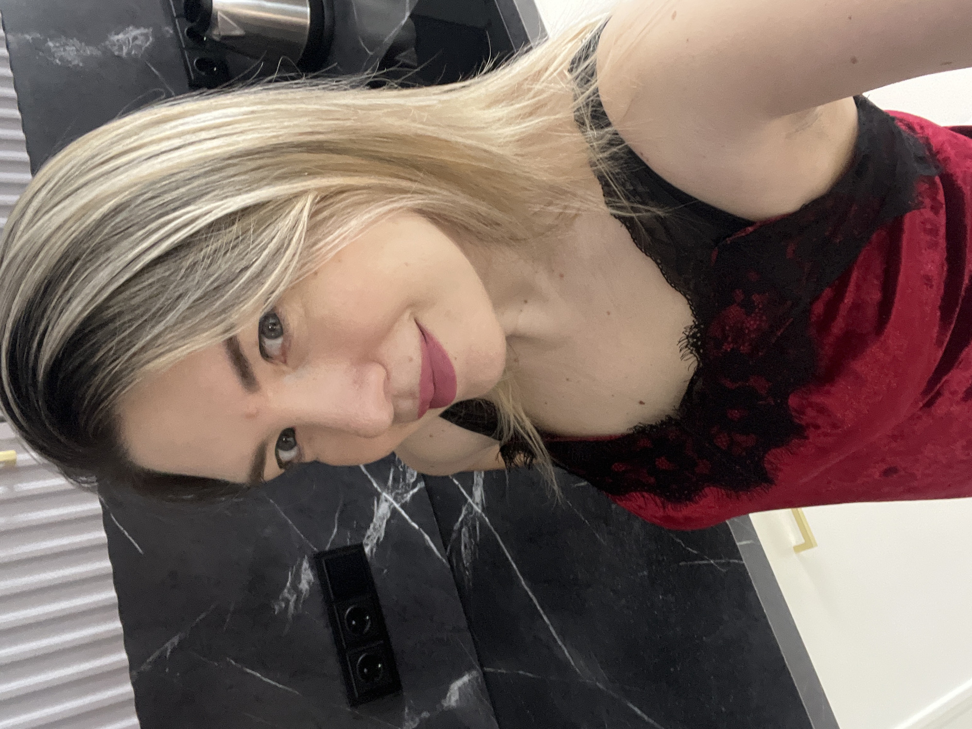 A woman with blonde hair and bright pink lipstick is taking a selfie in a kitchen. She is wearing a red velvet top with black lace trim. The kitchen has black marble countertops and white cabinets. The background includes electrical outlets and a kettle.