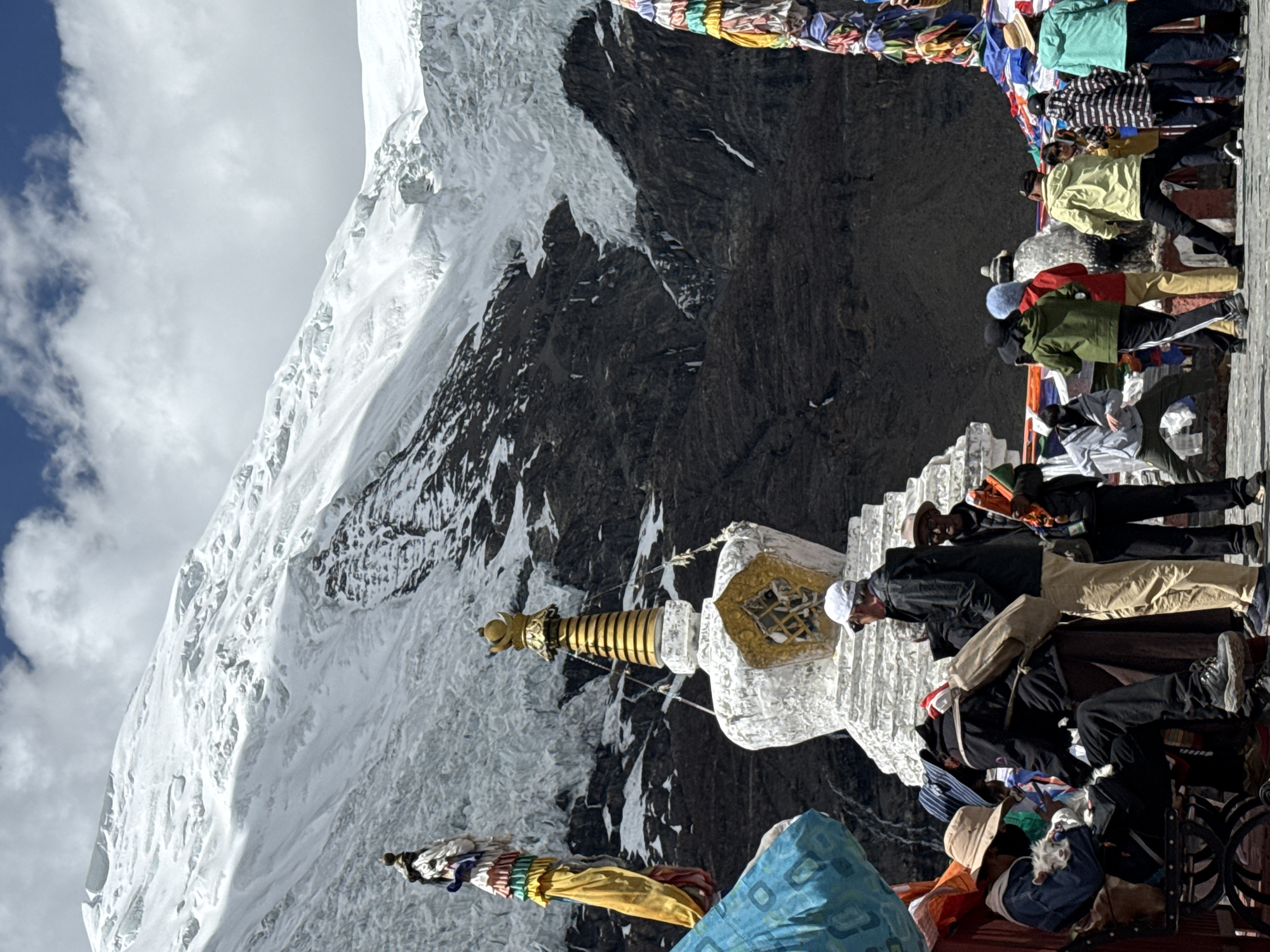 The image shows a view of a snowy mountain landscape in Gyantse County, China. In the foreground, a white and gold stupa (Buddhist shrine) stands prominently, adorned with prayer flags. A group of people is gathered on a structure near the stupa, with some sitting and others standing. The landscape features a combination of snow-covered peaks and dark, rocky terrain. The sky is partly cloudy.