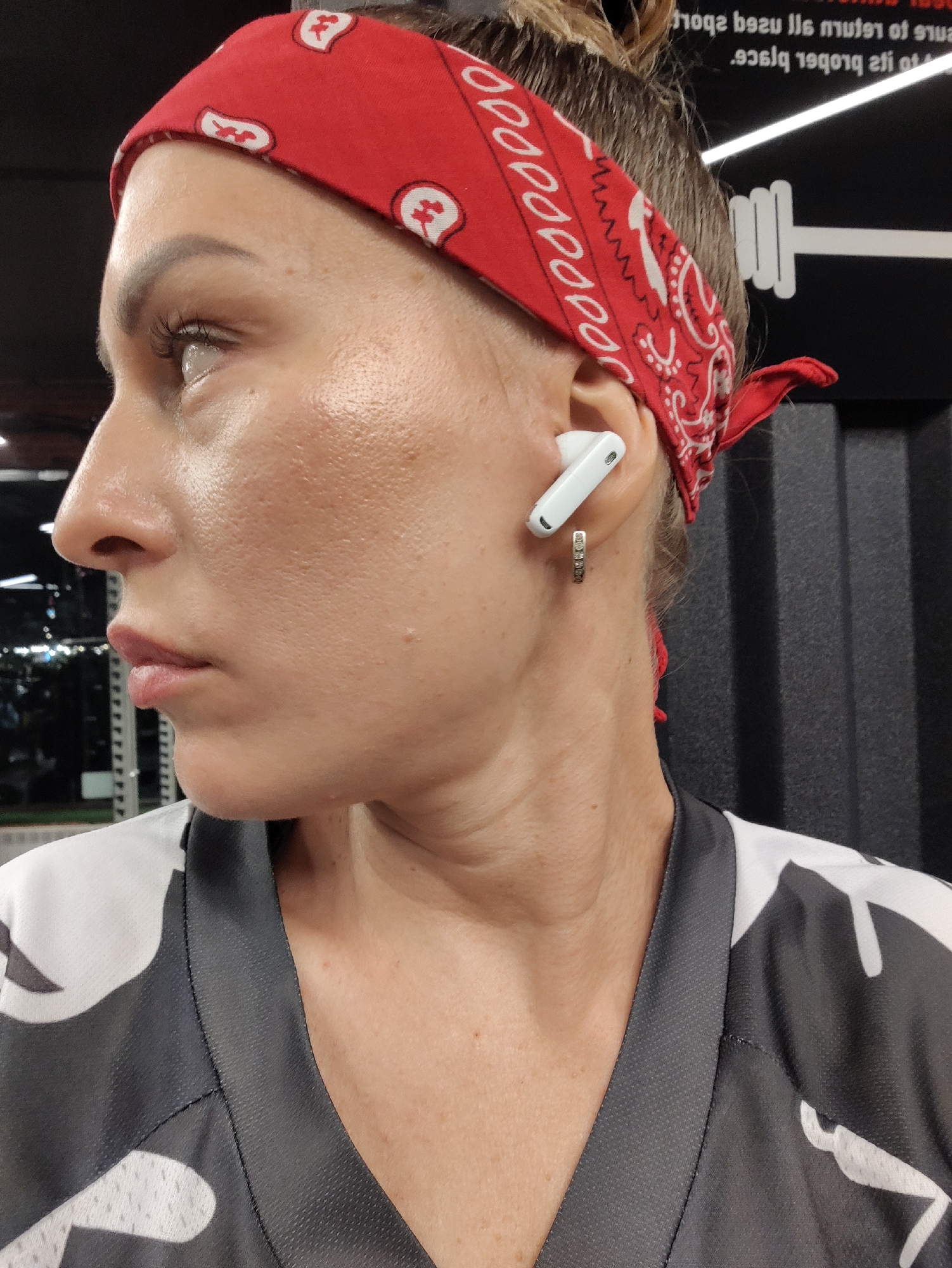 This is a medium shot, profile view of a woman in what appears to be a gym. The woman has blonde hair pulled back in a bun and is wearing a red bandana with a white paisley pattern. She is also wearing a white and gray patterned athletic top. She has a white earbud in her right ear and a small hoop earring. The background shows a black wall with a white barbell graphic and some text, which is difficult to read clearly due to the image quality and glare. The text appears to be in English and may be related to gym rules or hours. The lighting suggests it is indoors, likely during the day. The woman's skin is slightly shiny, possibly from sweat, indicating she may be in the middle of a workout. The overall mood seems to be focused and determined.