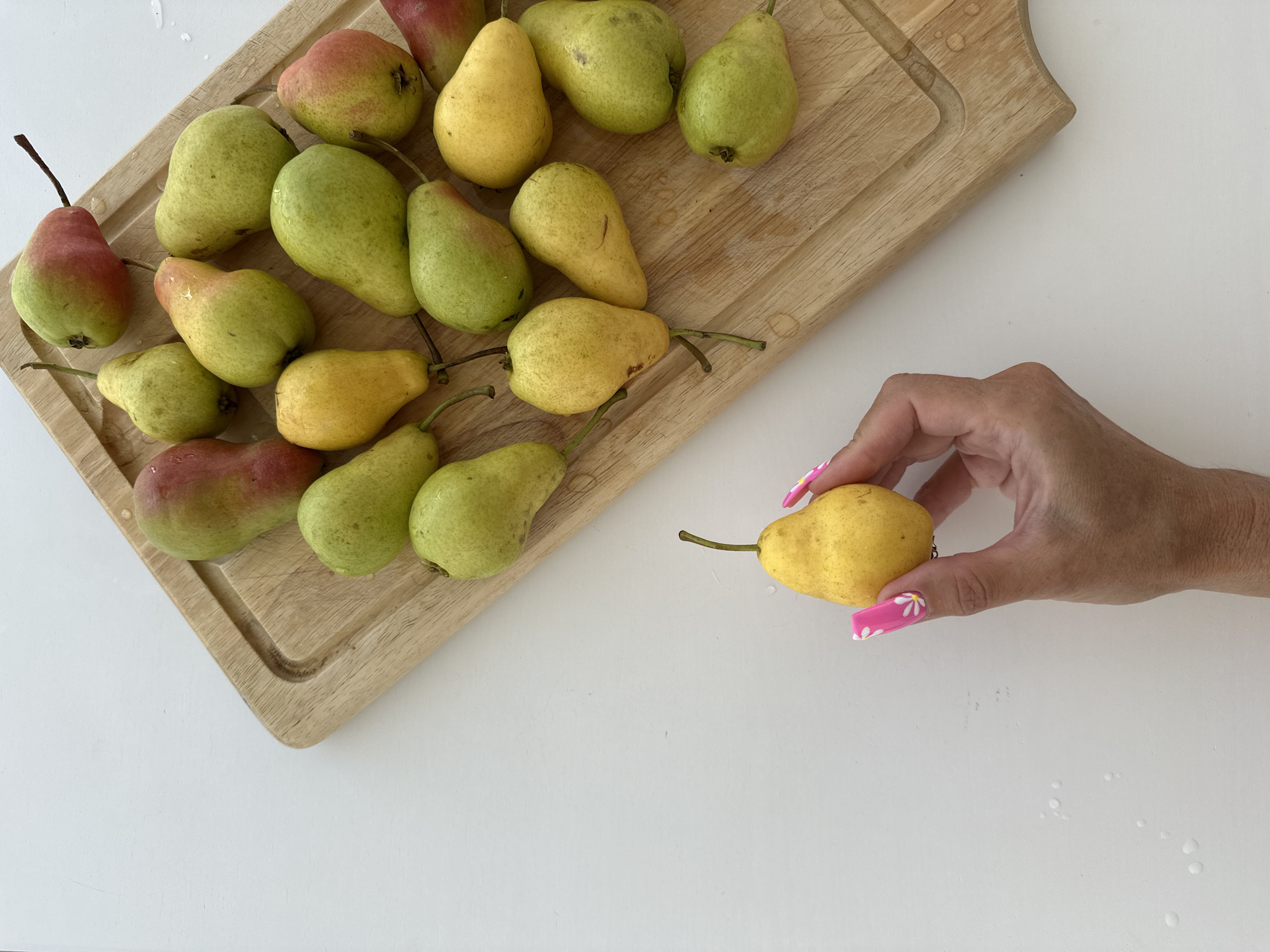 A close-up, overhead shot captures a wooden cutting board filled with a variety of pears. The pears are an assortment of yellow and green hues, with some exhibiting red blushes. A hand with brightly colored pink nails, adorned with white floral designs, is gently holding a single yellow pear in the lower left quadrant of the image. The pears are arranged on a light-colored surface, likely a countertop or table. The scene is clean and well-lit, with no visible signs of wear or damage to the pears or the cutting board. The overall impression is one of freshness and natural beauty. The image is not located in Tyumen, Russia.