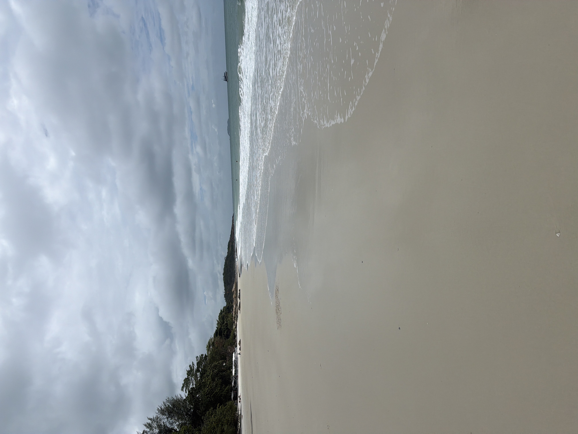 The image depicts a serene beach scene in Ao Hin Khok, Thailand, under a cloudy sky. The foreground is dominated by a wide expanse of pale, smooth sand, wet near the water's edge where gentle waves are breaking. The sea is a muted turquoise, with white foam marking the surf. In the distance, a small boat can be seen on the horizon. Along the left side of the frame, a line of lush green trees separates the beach from what appears to be a small settlement with white structures, possibly umbrellas or temporary shelters. A few scattered rocks are visible on the sand further down the beach. The overall atmosphere is calm and slightly overcast, suggesting a peaceful day at the coast.
