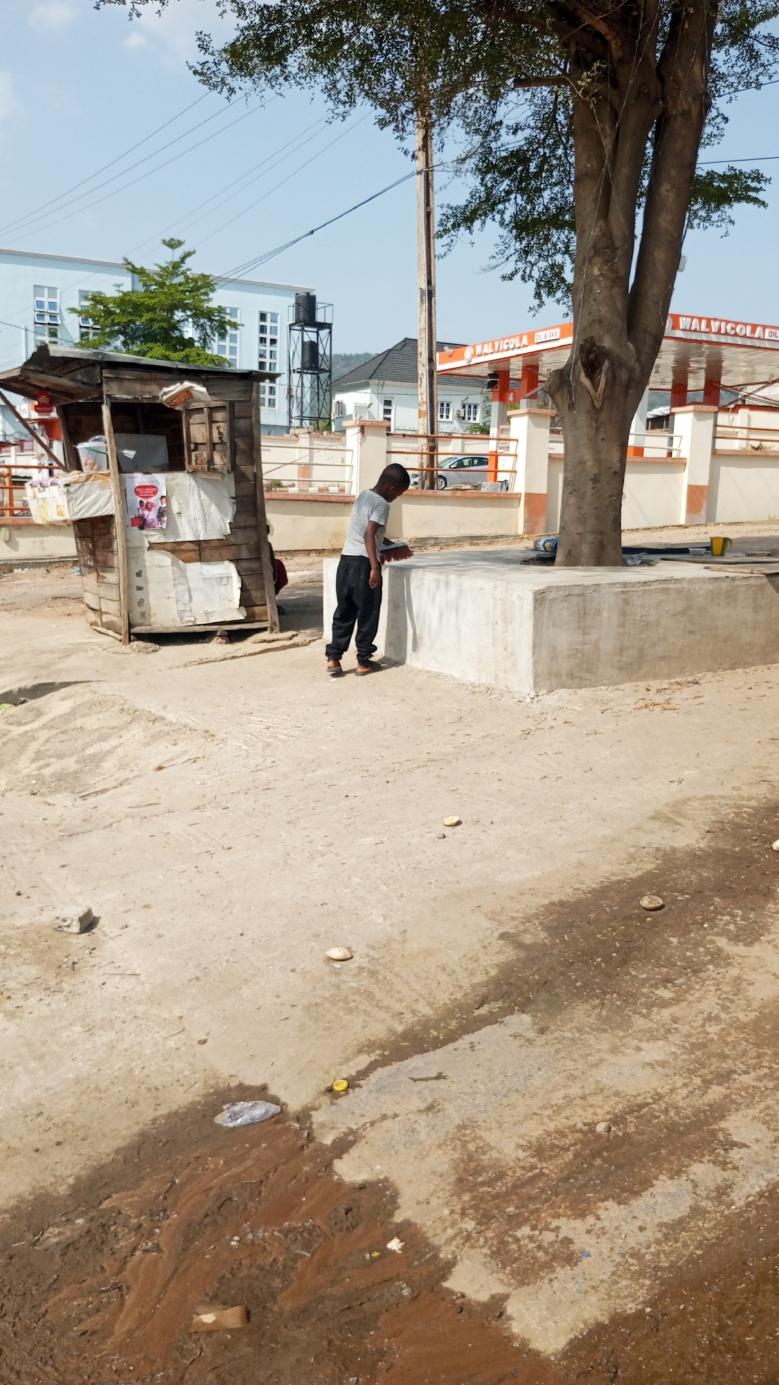 The image depicts a scene in Adankolo, Nigeria. A young person stands in front of a raised concrete structure that surrounds the base of a tree. They are wearing a light-colored t-shirt and dark pants, and appear to be sweeping the surface of the concrete with a brush. To the left of the person is a small, weathered wooden structure resembling a kiosk. In the background, buildings and a Walvicola gas station are visible. The ground is uneven and appears to be a mix of concrete and dirt, with some debris and a patch of muddy water. A utility pole stands near the tree.