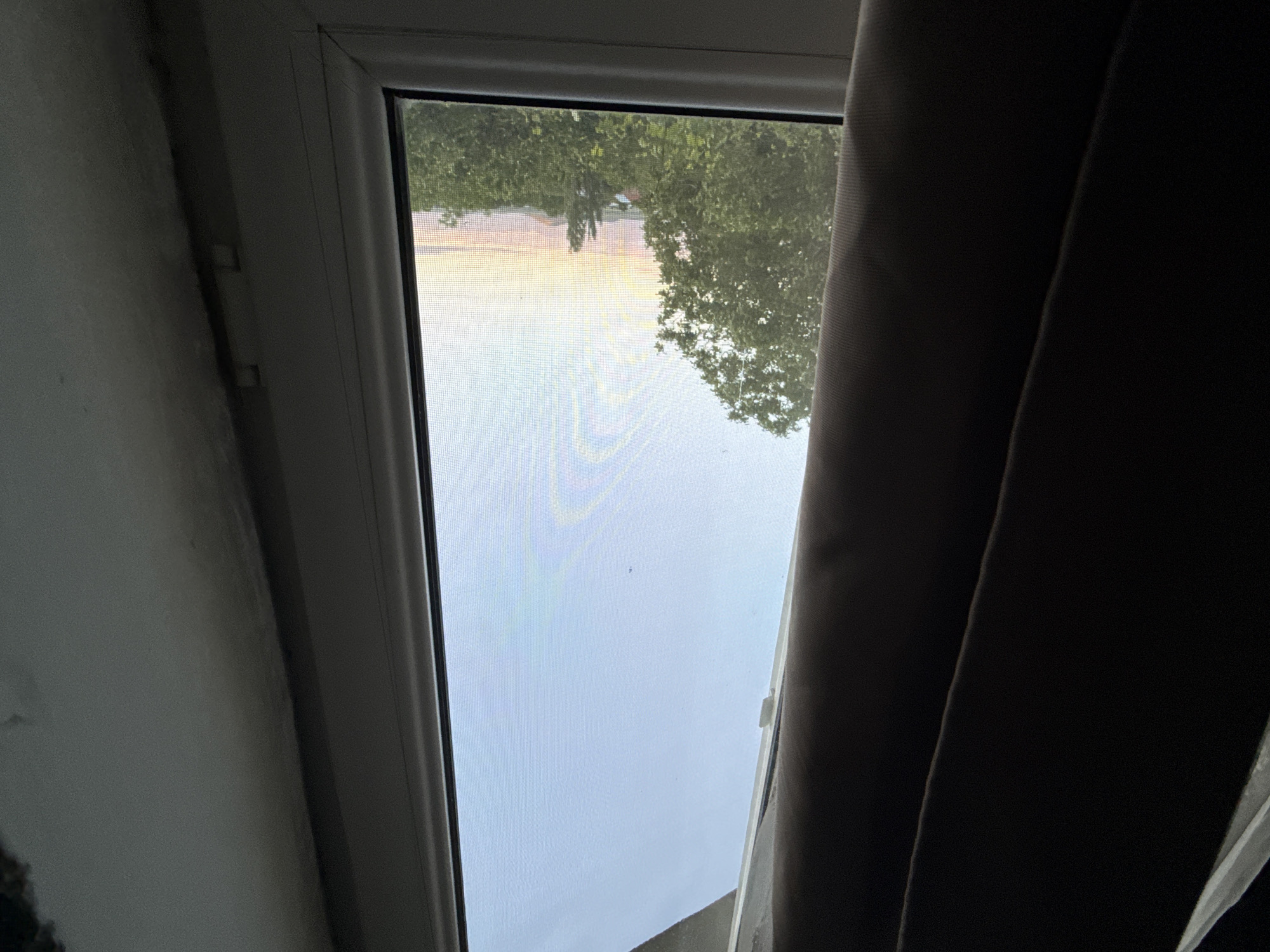 The image presents a view looking outwards from a window, framed by an interior setting. The primary subject is a white UPVC window frame with a visible mesh screen covering the glass, through which the outside world is seen.
Outside the window, a landscape of lush green trees is visible, suggesting a natural or suburban environment. Above the trees, the sky displays soft hues of pink, orange, and yellow along the horizon, transitioning to a lighter grey-blue further up, indicating either sunrise or sunset. The fine grid pattern of the window screen is prominent across the entire view of the sky and trees.
To the left of the window, a light-colored wall is partially visible, appearing somewhat grimy or discolored near the window frame. A vertical line or trim runs along the wall next to the window, with two small, rectangular, light-colored fixtures or clips attached to it. On the right side, a dark brown or charcoal-colored curtain hangs, partially drawn across the window, obscuring a portion of the view and casting a shadow.
The scene is static, with no activity or people depicted. The lighting suggests a transitional period of the day, likely early morning or late afternoon/evening, characterized by the soft, colored light on the horizon and the dimmer interior. There is no visible text in the image.