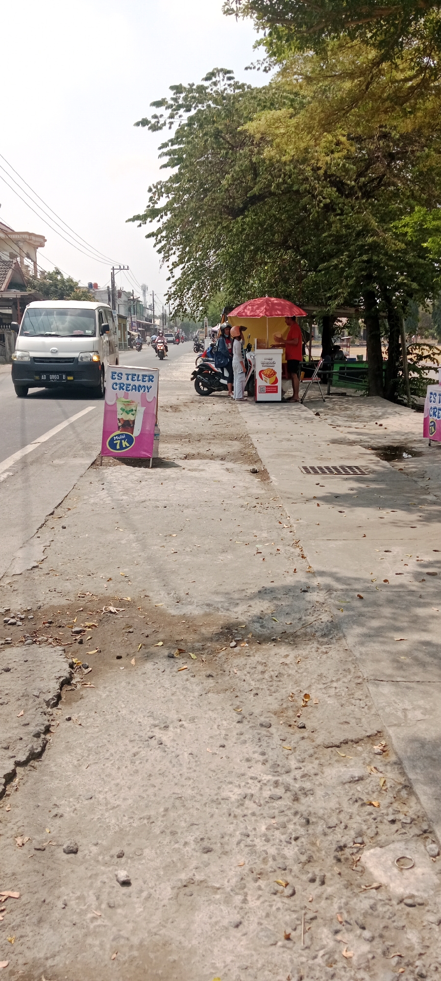 On a sunny day in Maguwoharjo, Indonesia, a white van with license plate AB 8860 B is seen driving on the left side of a two-lane road. On the sidewalk to the right, an "Es Teler Creamy" advertisement stands prominently. Further down the sidewalk, a food cart is set up under a red umbrella. Two individuals, one wearing a red shirt and the other in light-colored clothing, are interacting at the cart, with a motorcycle parked nearby. The environment appears to be a roadside, with buildings and trees lining the street. The sidewalk itself shows signs of wear and tear, with cracks and scattered leaves.