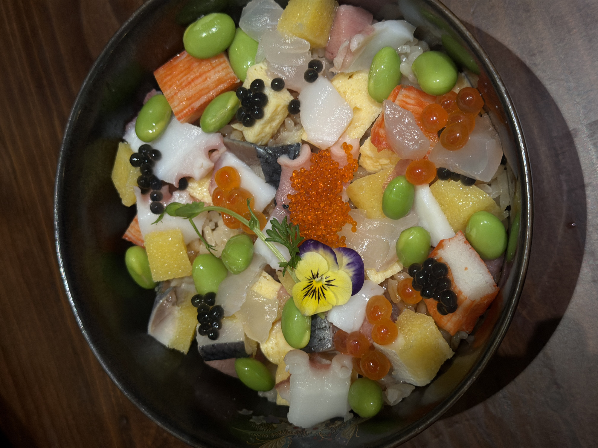 A top-down, close-up shot reveals a bowl of chirashizushi, a Japanese rice bowl dish, filled with an assortment of ingredients. Visible are pieces of fish, octopus, crab sticks, cooked egg, edamame beans, various types of roe (orange and black), and a yellow flower garnish. The bowl is dark in color and set on a wooden table. There are no people present in the image. The location in the city of Đà Nẵng, Vietnam is unknown.