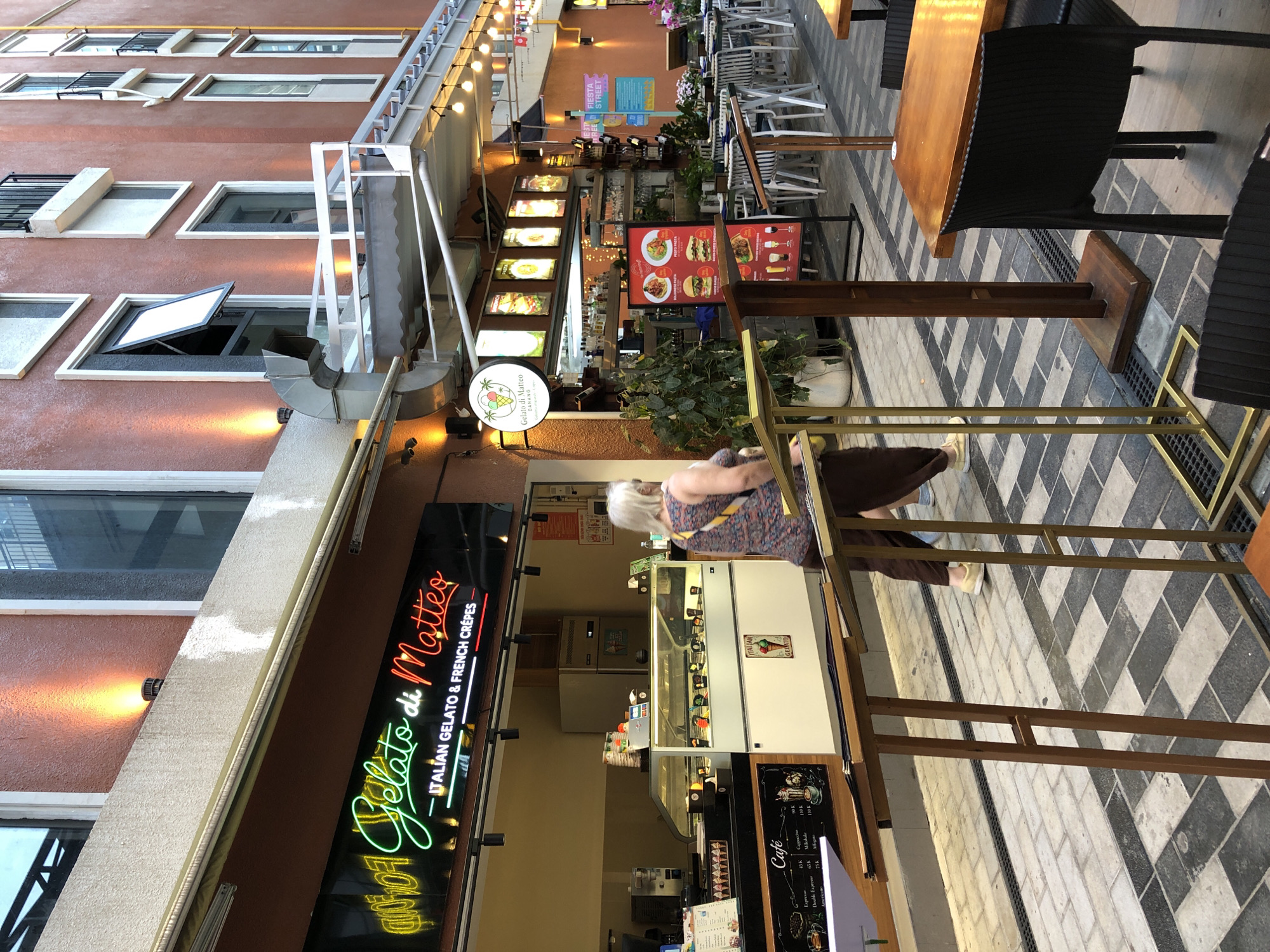 The image depicts a street-level view of an outdoor establishment in Đà Nẵng, Vietnam. A neon sign above the storefront reads "Gelato di Matteo - ITALIAN GELATO & FRENCH CREPES." The establishment appears to be a gelato shop and cafe, with a display case filled with colorful gelato visible inside. To the right of the entrance, tables and chairs are set up for outdoor dining, with some patrons already seated. Further down the street, more tables and umbrellas suggest a bustling dining area. A woman with blonde hair is walking away from the camera, wearing a patterned top and brown pants. The architecture features a reddish-orange facade with multiple windows and balconies. The pavement is a decorative pattern of grey and black tiles. The overall scene conveys a lively atmosphere in a commercial district.