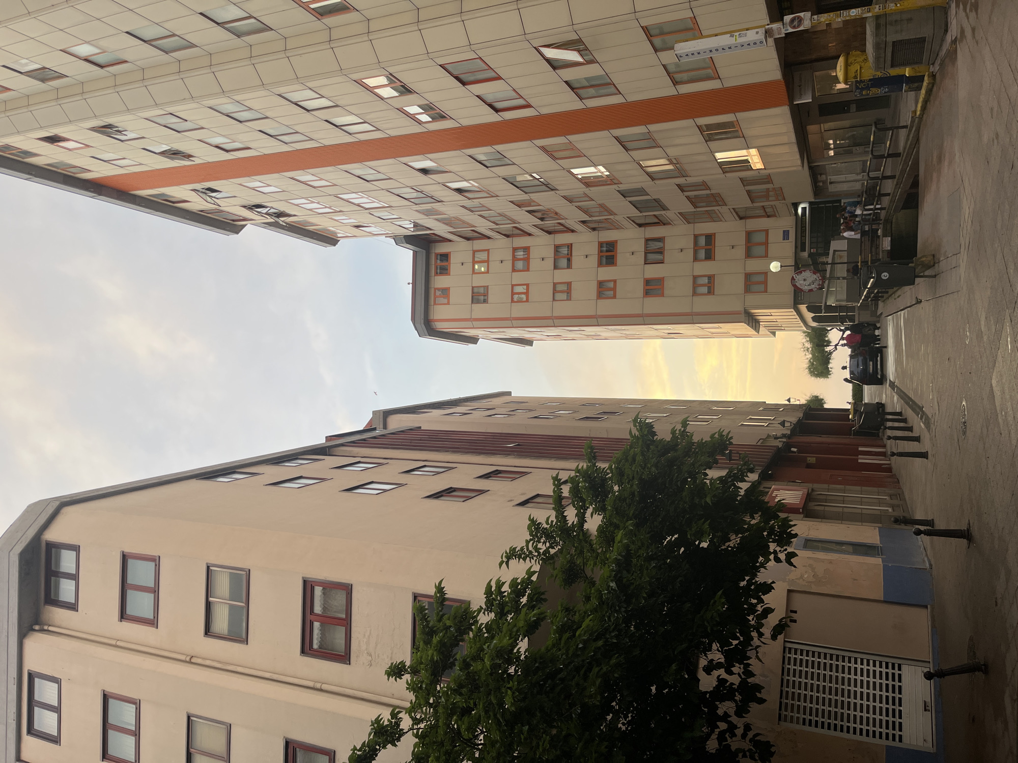 The image shows a narrow street in A Coruña, Spain, framed by tall buildings. The buildings are beige with rows of windows, some with red frames. The sky is visible between the buildings, appearing light yellow at the horizon, suggesting sunset or sunrise. A tree with green foliage is present in the foreground on the left. The street is paved and appears to lead to a slightly brighter area further down. There are bollards along the sidewalk on the right side of the street. People are partially visible at the end of the street. The image is angled upwards, emphasizing the height of the buildings. A street sign is visible on one of the buildings. There is a parked car and other vehicular traffic at the end of the street. The overall scene is urban and suggests a residential or mixed-use area.