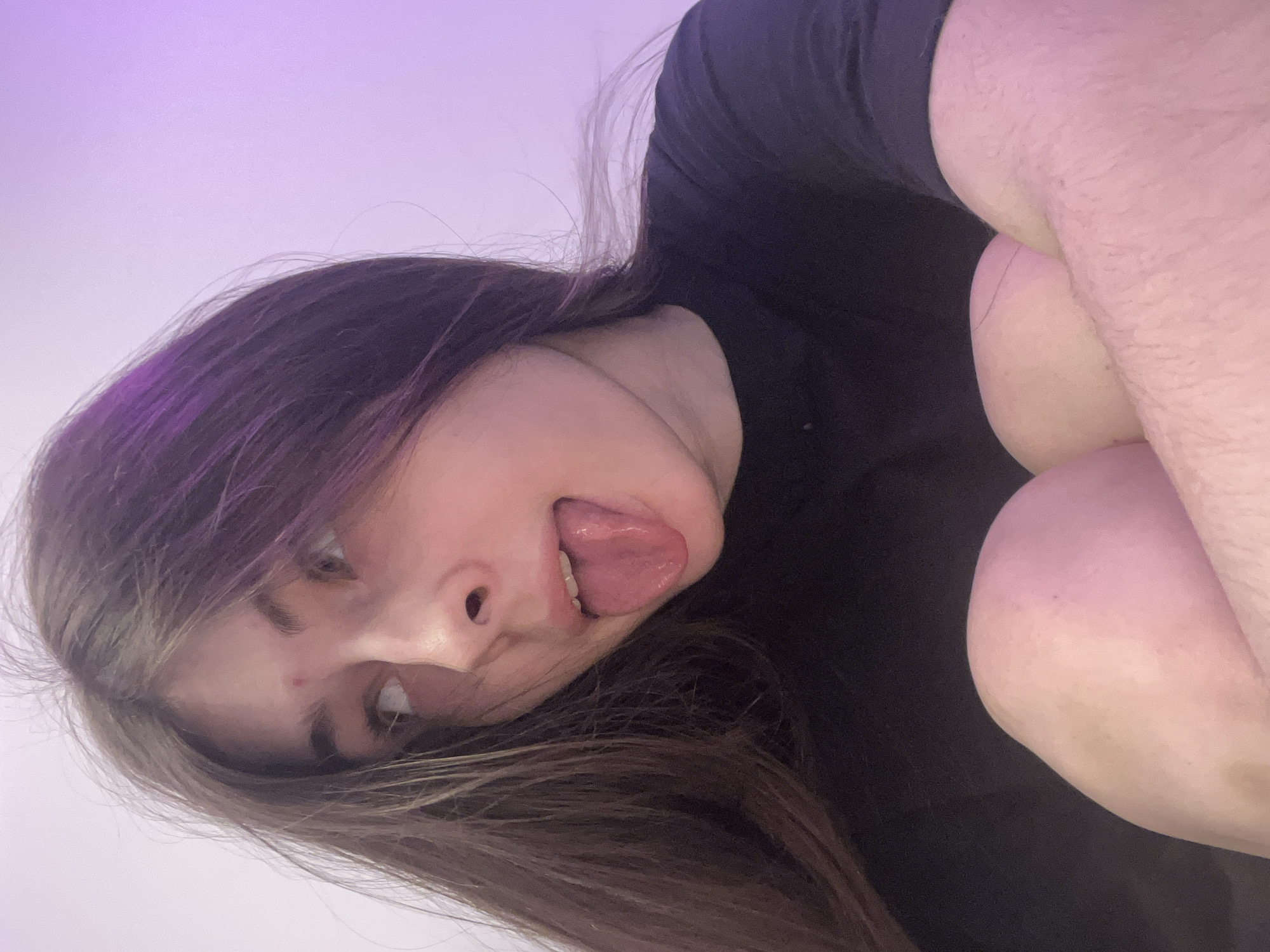 A young person is positioned in the frame with their head and upper body visible. They have long brown hair with some purple streaks and are sticking their tongue out. They are wearing a black shirt. Part of their arm is visible, bent at the elbow and held close to the body. The background is a blurry white and purple gradient. There are no indications of Saint Petersburg, Russia.