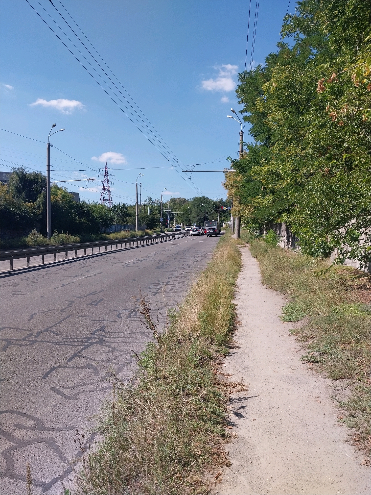 The image shows a sunny day on a paved road with a dirt path running alongside it in Dnipro, Ukraine. The road is bordered by a metal guardrail on the left and a concrete wall with trees and vegetation on the right. Overhead, multiple power lines and trolleybus wires crisscross the sky, supported by poles. In the distance, a tall red electrical pylon is visible. Traffic is present on the road, with cars and a bus. The ground on the road shows markings. The path is narrow and appears unpaved, with grass and dry vegetation growing along its edges. The sky is blue with scattered white clouds.