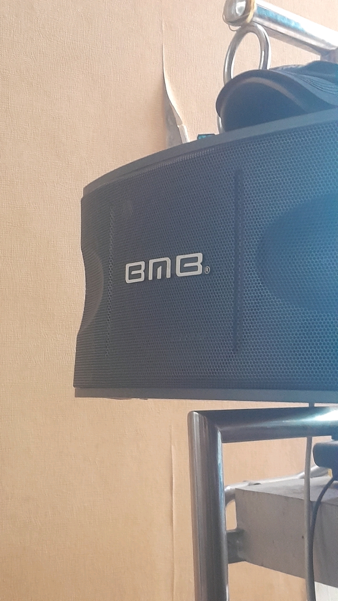 The image shows a dark grey speaker with the "BMB" logo embossed on it. The speaker appears to be suspended or mounted on a metal rack. In the background, there is a light brown wall with some peeling wallpaper. There are also hints of other objects, possibly part of a sound system or furniture, below and to the right of the speaker. The overall scene suggests an indoor setting, possibly a room or a shop, and the presence of a sound system might indicate a place of entertainment or a retail environment. There is no discernible indication of the specific location within Madiun, Indonesia.