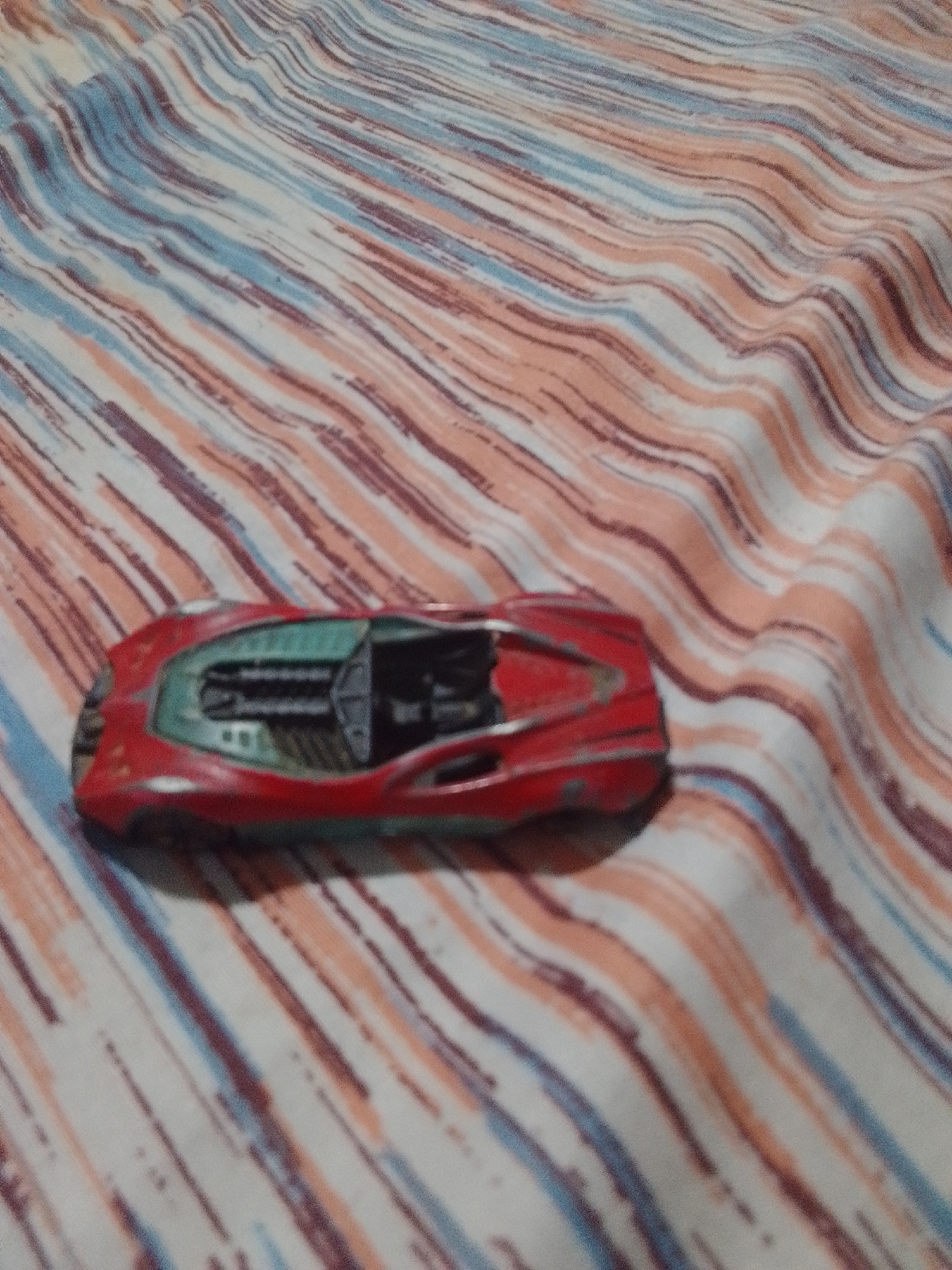 A red toy car with a green interior is positioned diagonally on a striped fabric surface. The fabric has alternating stripes of white, peach, and dark blue. The toy car is in focus, while the fabric is slightly blurred. There are no people or any other objects visible in the image. The location in the city of La Tagua, Venezuela cannot be determined from the image.