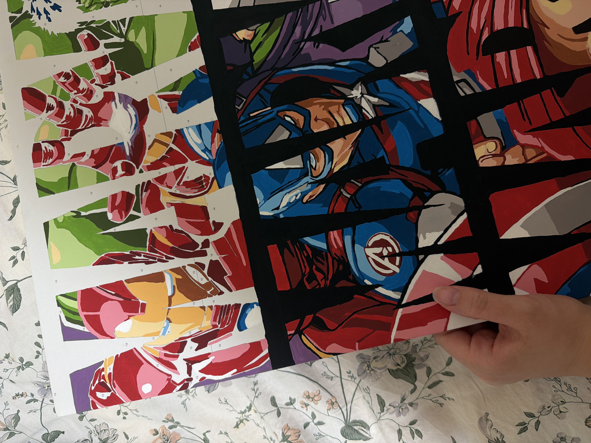 The image shows a partially completed paint-by-numbers canvas featuring three Marvel superheroes: Iron Man, Captain America, and what appears to be another character on the right (possibly another Avengers member). The canvas is divided into color-coded sections, some of which have been painted. The painted sections reveal the characters in a dynamic, comic-book style. The canvas is held at the edge by a hand and rests on a bedspread with a floral pattern.
There is no information in the image about it being in Izhevsk, Russia.