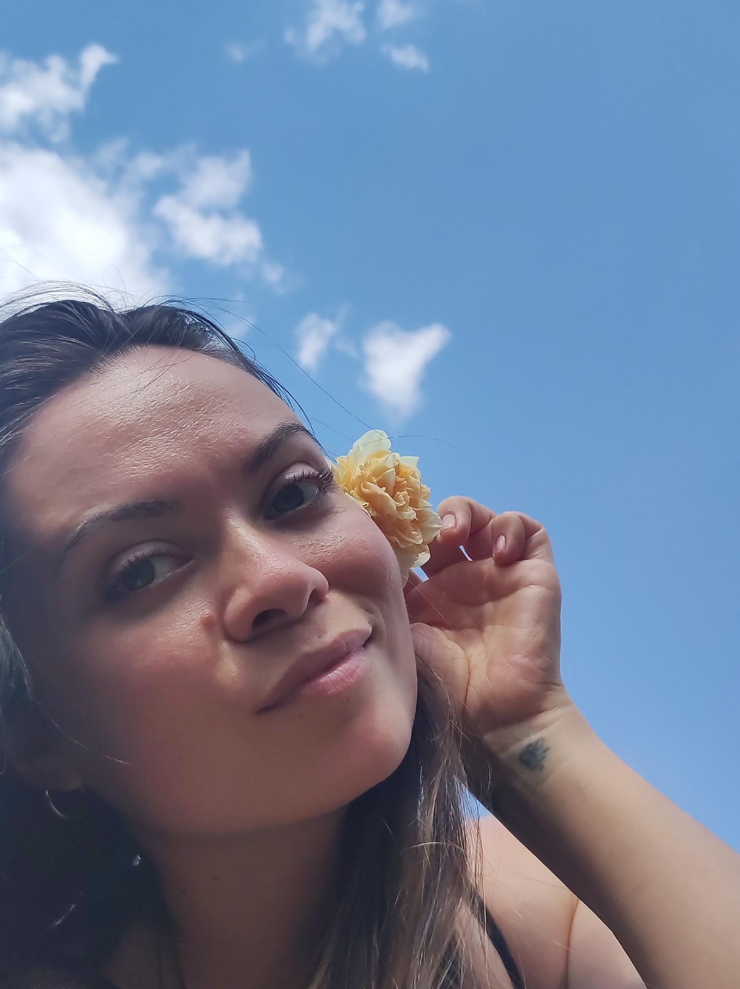 A young woman poses for a selfie against a blue sky with scattered white clouds. She is holding a pale yellow flower near her right ear. Her expression is pleasant. The woman has brown hair and fair skin. A tattoo is visible on her left wrist. No contextual details indicate a specific location; Фоки, Russia, cannot be confirmed from the image alone.