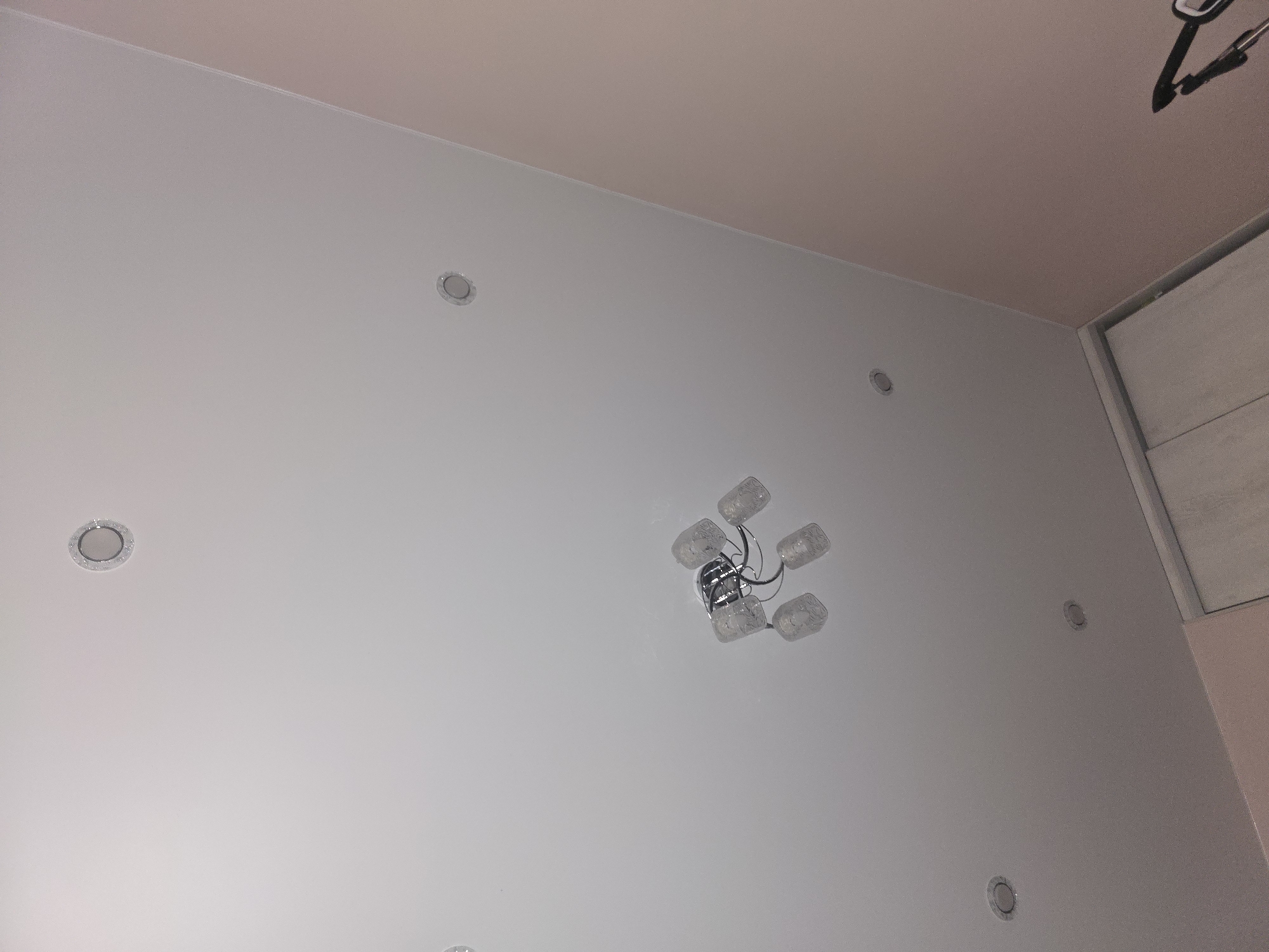 The image shows a modern ceiling with several recessed spotlights and a chandelier. The chandelier is centrally located and has five glass shades. The ceiling transitions to a pink wall on the right side of the image, and a portion of a wardrobe with white doors is visible at the bottom. The scene is an indoor residential space, consistent with a room in Ulan-Ude, Russia, characterized by contemporary interior design. There are no people or visible actions taking place.