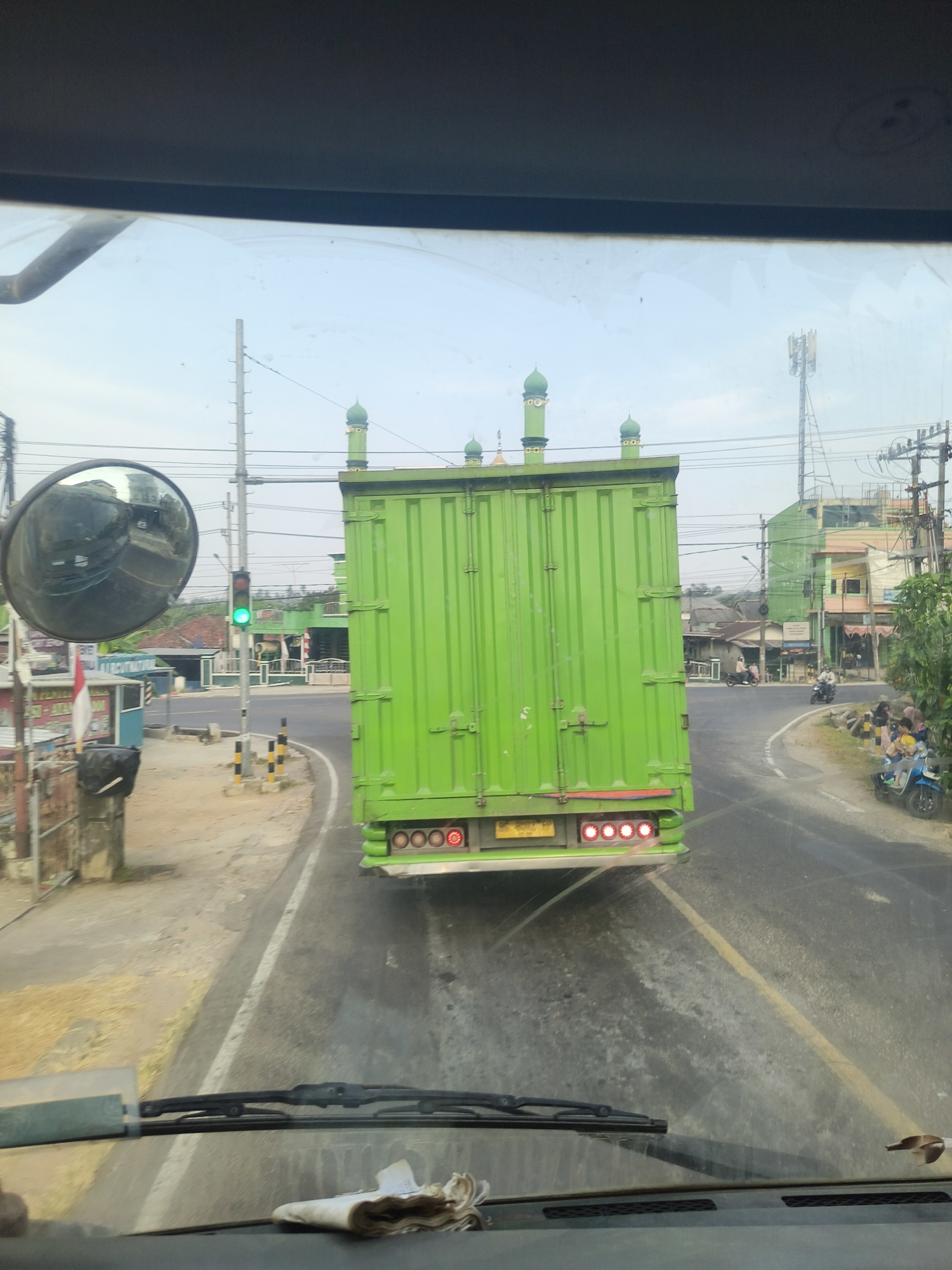 The image is taken from inside a vehicle, looking out through the windshield at a bright, overcast sky. In the center of the frame, a large, lime-green truck is stopped at an intersection. The truck's rear doors are closed, and it has a cargo box with several green dome-shaped structures on top, resembling minarets. The truck's rear lights are illuminated.
To the left of the truck, a traffic light shows a green signal. A large, round side mirror from the vehicle in front is visible, reflecting the surrounding scene. The intersection itself appears to be paved with asphalt and has some road markings. Sidewalks and the sides of the road are visible, with some structures and vegetation.
In the background, on the right side of the truck, there are buildings, including a brightly colored, multi-story structure with signs. A cell tower is also visible in the distance. Several people are seen on the right side of the road, and some motorcycles are present. A red and white Indonesian flag is attached to a pole on the left side of the road. The overall environment suggests a suburban or semi-urban setting in Indonesia. The license plate on the truck appears to be from Indonesia.