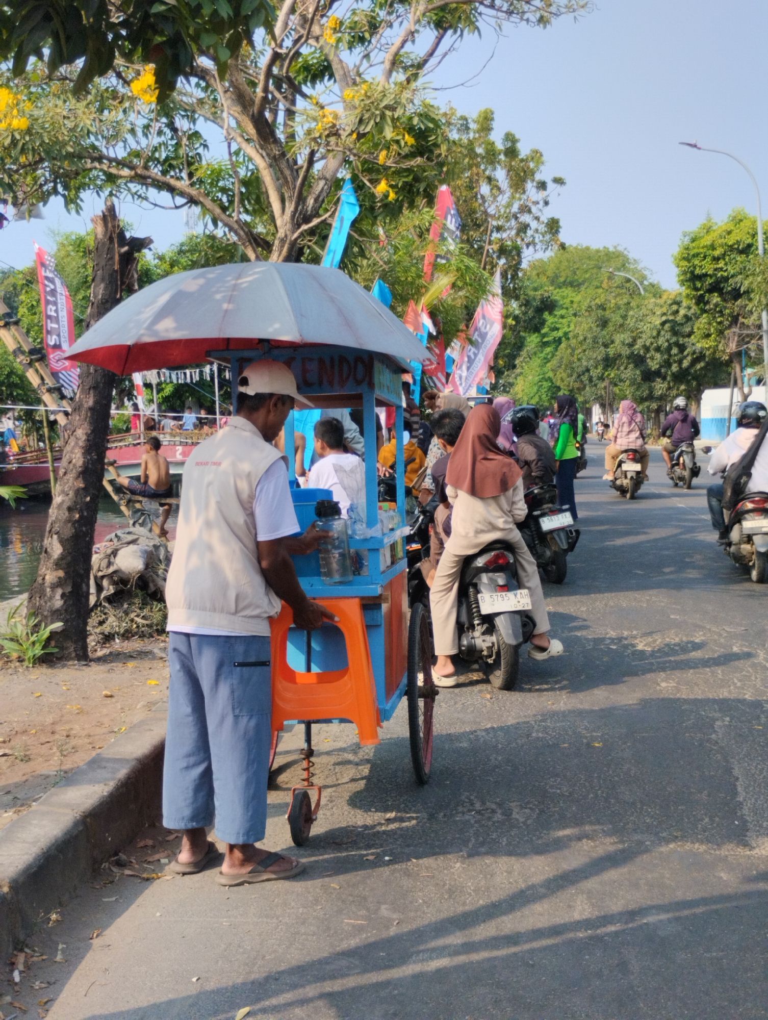 A street vendor with a cart stands on the side of a road in Bekasi, Indonesia. The vendor, a man wearing a light-colored vest over a white t-shirt and blue pants, holds a large plastic water bottle. His cart, a small structure with a red and blue frame and an orange plastic seat, is equipped with an umbrella for shade. In the background, several people ride motorcycles on the paved road, some wearing helmets and others headscarves. Trees line the roadside, and flags of various colors are visible, suggesting a public event or campaign. A canal or waterway runs alongside the road, with a person sitting on its edge in the mid-ground. The sky is clear and blue, indicating a sunny day.