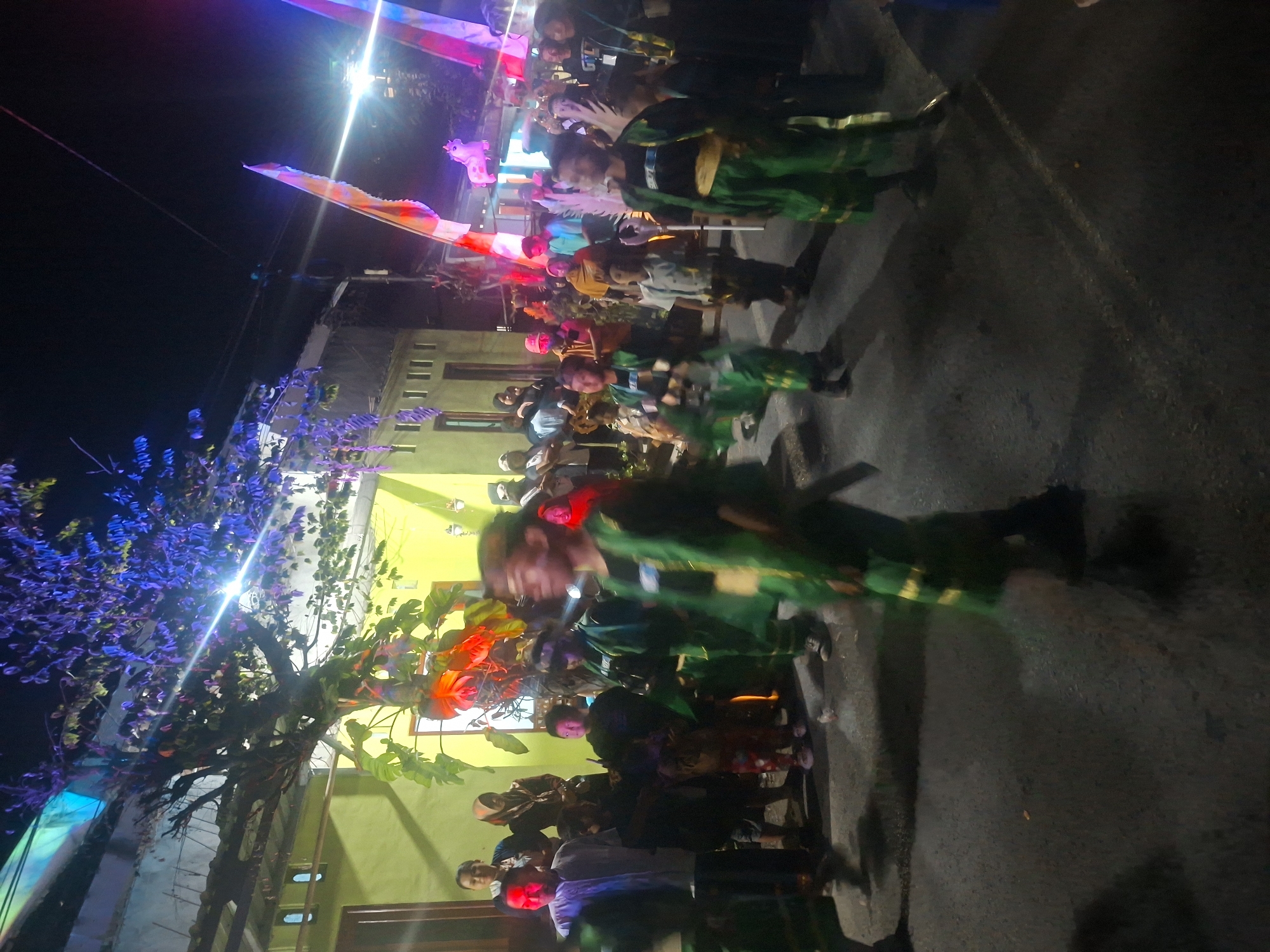 A nighttime outdoor street scene in Tongaswetan, Indonesia, depicts a cultural parade or performance. A group of children in matching green costumes, featuring gold accents and sashes, are marching in the foreground. Their costumes include vests worn over black shirts and matching pants. Some of the children are blurred due to motion. Behind them, a crowd of spectators watches from the roadside. Buildings with yellow walls and tiled roofs line the street. The scene is illuminated by colorful lights, including vibrant purple lights reflecting on a tree's foliage and pink and blue lights from banners or decorations. Flags are visible, one of which has a unicorn figure on it. The ground is paved with asphalt, and some debris is scattered on it. The overall atmosphere suggests a festive community event.