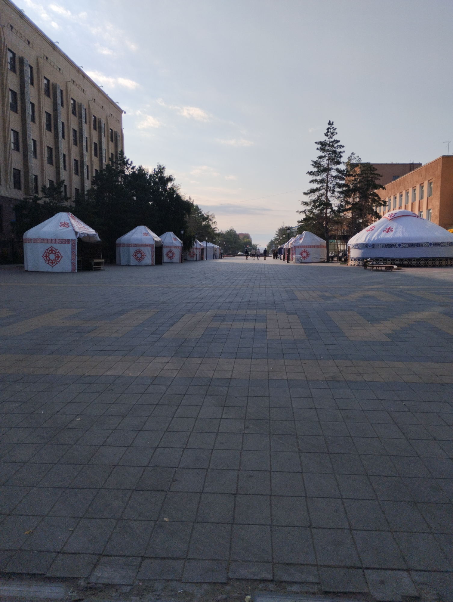 A wide, paved pedestrian area is lined with several yurts, also known as gers, which are traditional nomadic dwellings. The yurts are decorated with red and white patterns. In the background, there are buildings and trees, suggesting an urban environment. The sky is clear with a few wispy clouds, and the light indicates it is either early morning or late afternoon. The pavement is made of dark gray and yellow square tiles, arranged in a grid pattern. Some people are visible walking in the distance, appearing as small figures on the pedestrian area. The scene is likely in Kostanay, Kazakhstan, given the presence of these structures often used for cultural events or displays in the region.