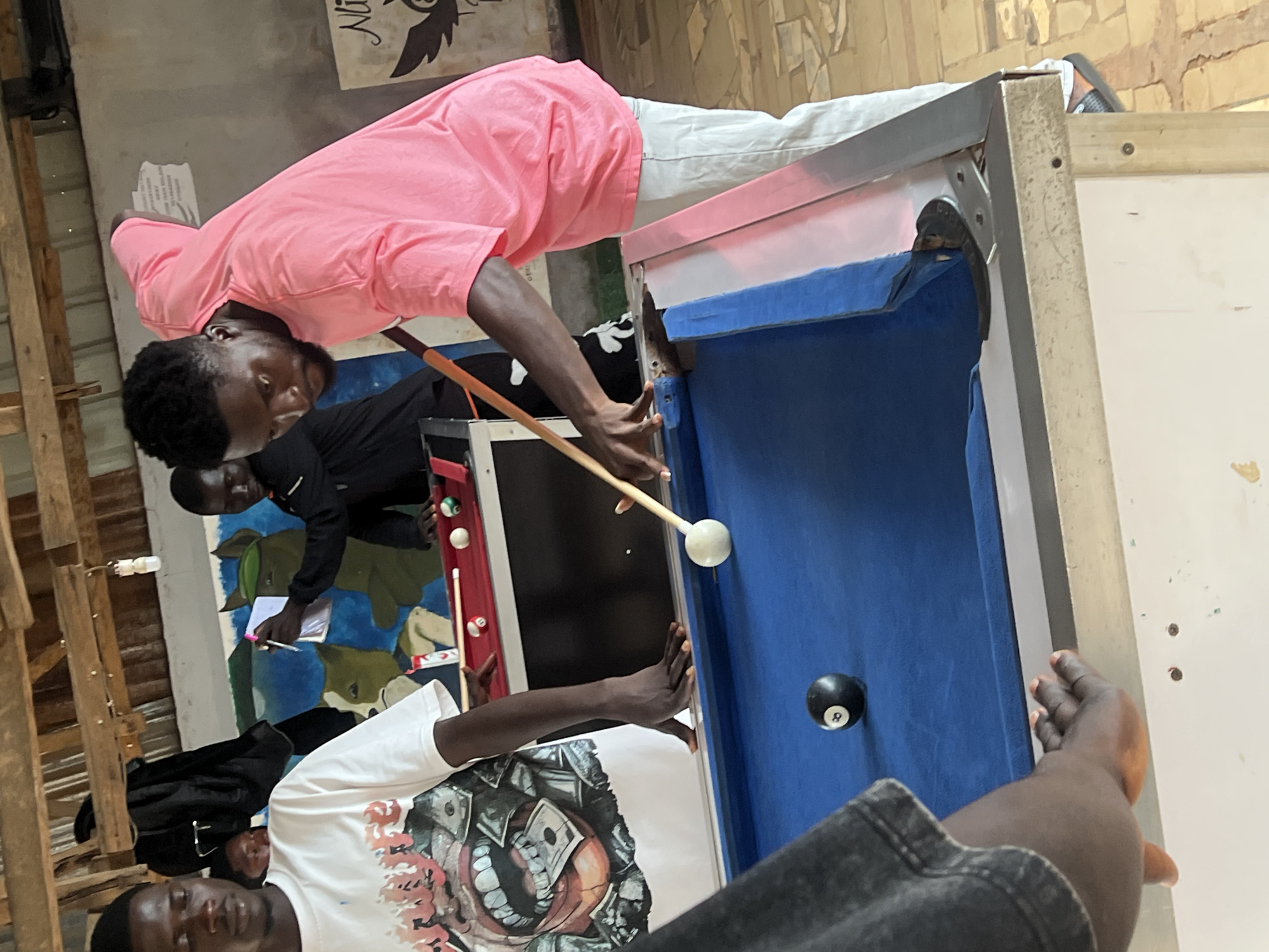 The image depicts an indoor scene with a pool table as the central focus. Three young men are visible. One man, wearing a pink t-shirt and light-colored pants, is actively playing pool, aiming the cue stick at a white ball. Another man, in a white t-shirt with a graphic print, is standing to the left, observing the game. A third man, wearing a black jacket, is in the background, holding a notepad and pen. The pool table has a blue felt surface and a red bumper. Several pool balls are on the table and in the corner pocket. The environment appears to be a casual gathering space, possibly a bar or recreational center, with wooden beams visible on the ceiling and a mural painted on the back wall. The scene is likely located in Durumi, Nigeria, given the provided context, though this cannot be definitively confirmed from the visual information alone.
