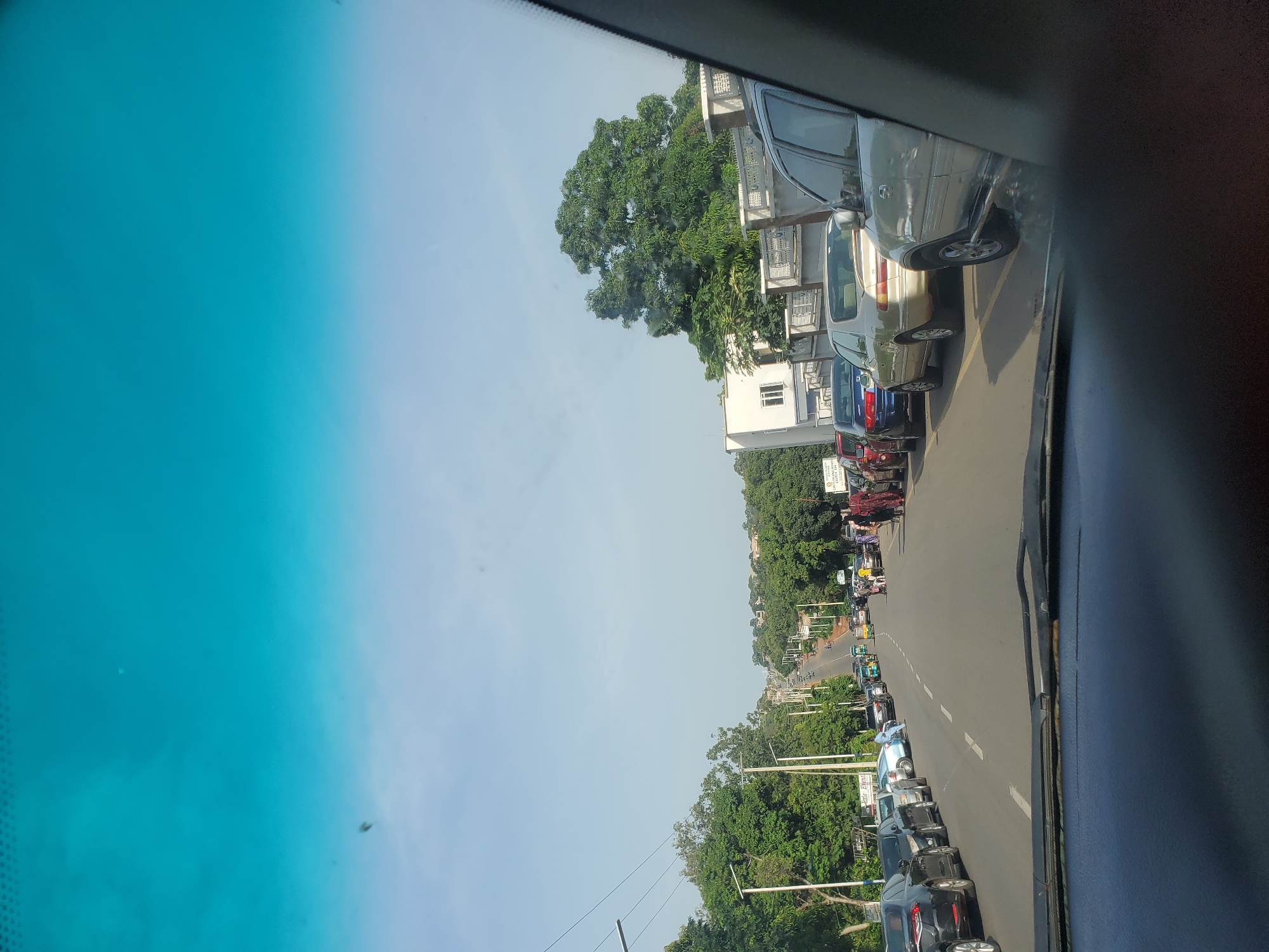The image depicts a street scene in Jos, Nigeria, viewed from inside a vehicle. The road is lined with parked cars and several vehicles are moving in traffic, indicating a busy urban environment. Tall green trees flank the sides of the road, and some buildings are visible in the background. The sky is a clear blue with a hint of clouds. The overall impression is of a typical day in a Nigerian city with vehicular activity and lush vegetation.