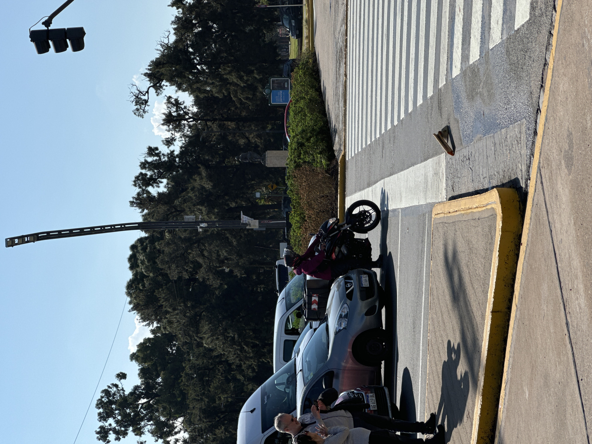 The image depicts a street scene in Buenos Aires, Argentina. On the right is a crosswalk with white stripes. Along the curb, marked with a yellow line, are parked vehicles. A motorcycle is stopped near the crosswalk. People are visible near the parked cars. Trees and foliage are present along the street. Above, a traffic light hangs from a pole against a blue sky. License plates visible on parked cars indicate Argentine registration.