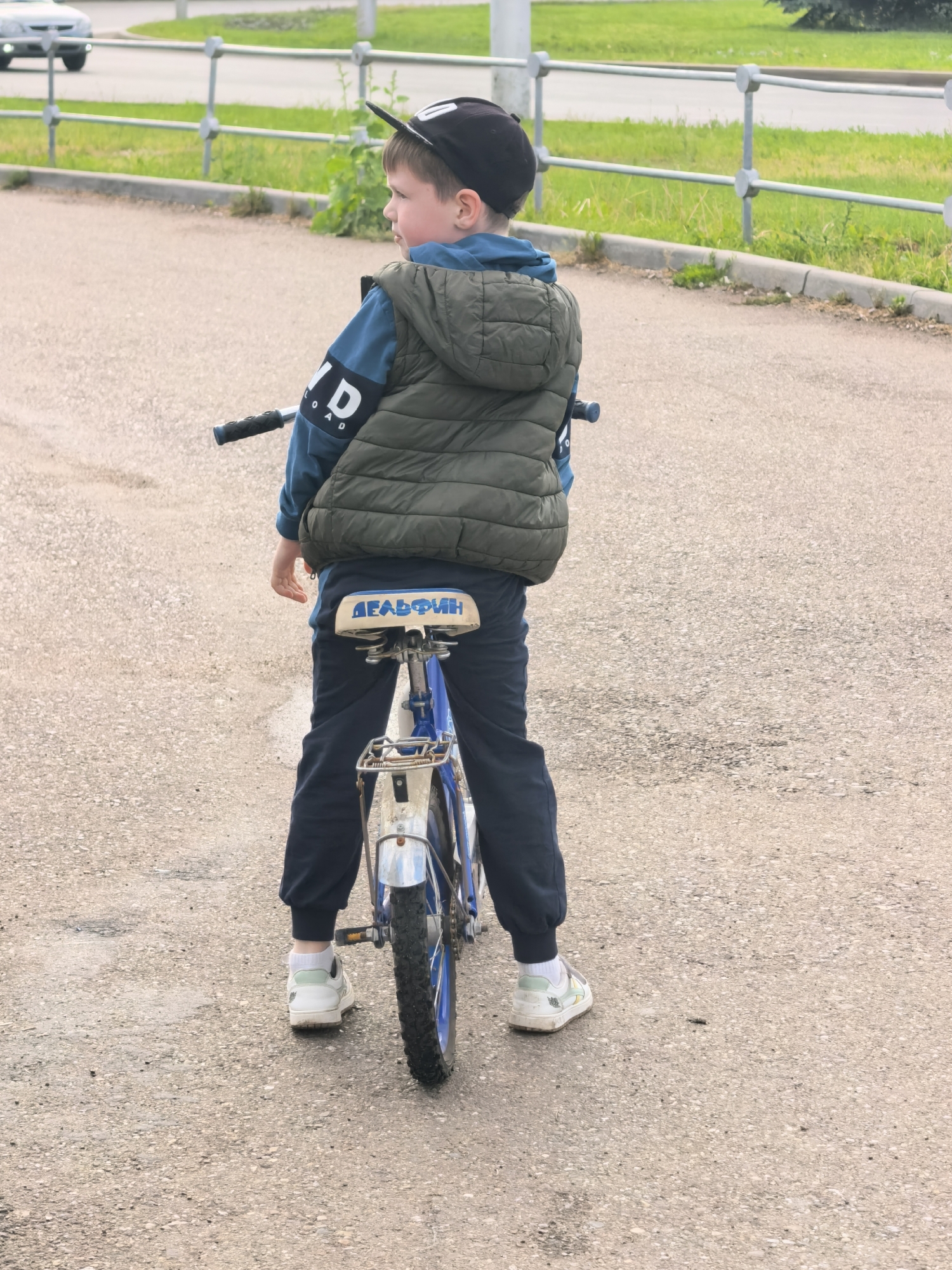 A young boy wearing a black cap, a green quilted vest over a blue sweatshirt with the word "ROAD" on the sleeve, and dark blue sweatpants stands behind a child's blue bicycle on a paved surface. He is facing away from the viewer, looking to the left. The bicycle seat has the word "ДЕЛЬФИН" (Dolphin) written on it. The boy is wearing white sneakers with a hint of green on the heel tab. In the background, there is a silver metal railing and green vegetation. A light-colored car is visible in the upper left corner. The scene appears to be set in a public area in Sterlitamak, Russia, based on the text on the bicycle seat.