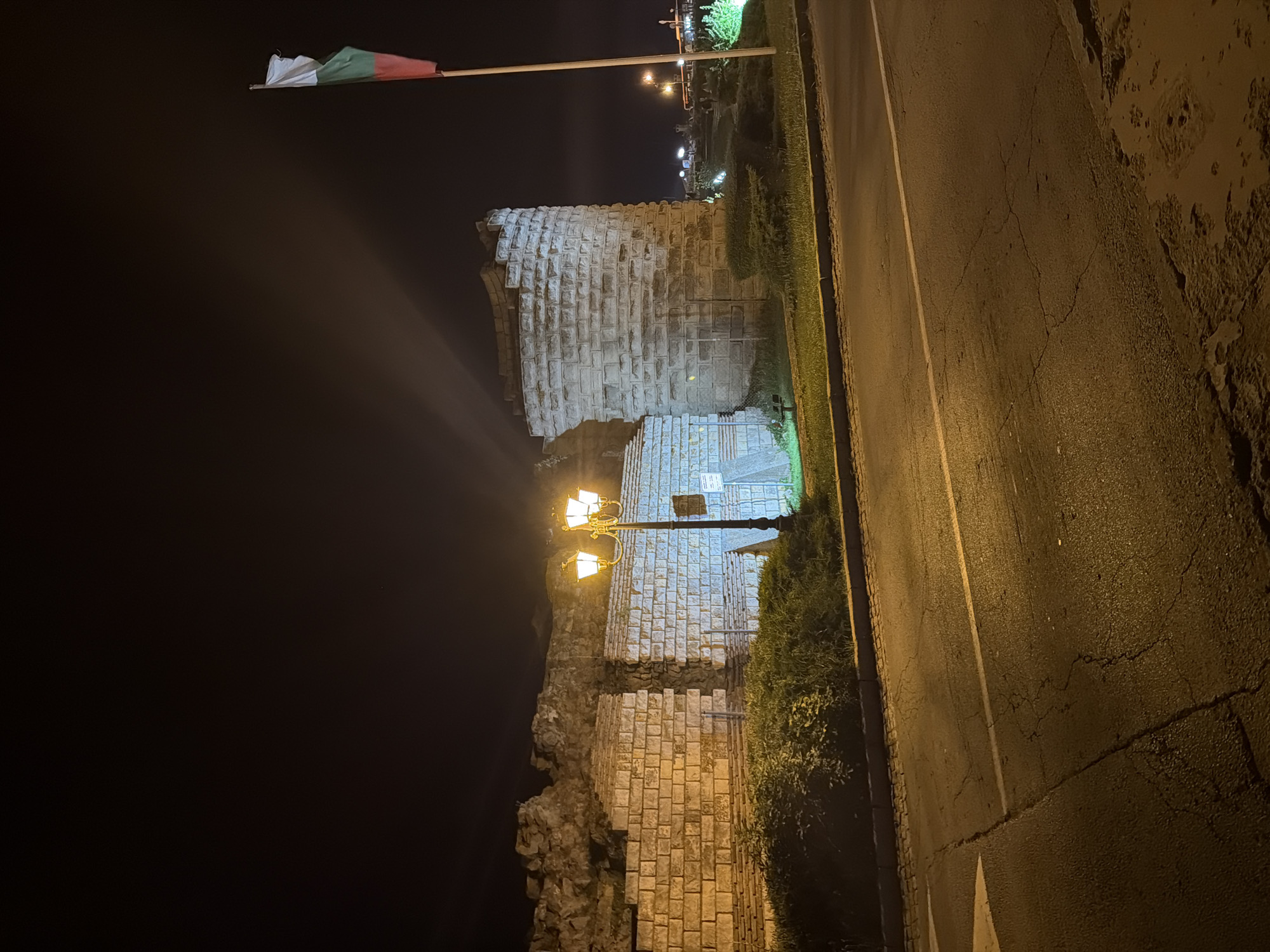 In Nesebar, Bulgaria, at night, a Bulgarian flag flutters from a flagpole next to a partially preserved ancient brick and stone fortress wall. The fortress wall, constructed from large, rough-hewn stones and smaller bricks, appears weathered and ancient, with sections showing signs of damage and decay. A distinct circular tower, also made of brick and stone, stands prominently to the right of the wall. The scene is illuminated by a tall, ornate streetlamp with four glowing bulbs positioned in front of the wall, casting a warm, yellow light. The area around the fortress is covered in dense green bushes and grass, bordering a paved road in the foreground. The road is asphalt, showing cracks and wear, with a white line marking a lane. The night sky is dark, with faint light beams visible emanating from behind the fortress. In the distance, beyond the fortress and a line of bushes, city lights are visible, suggesting the presence of other structures and activity in the vicinity. The overall impression is one of historical significance juxtaposed with modern infrastructure and nighttime illumination.