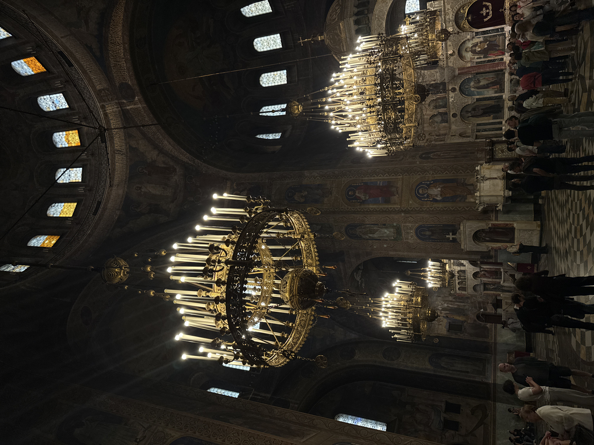 The image depicts the interior of a large church, likely located in Unknown, Unknown. The perspective is from below, looking up at ornate chandeliers hanging from the high ceiling. The chandeliers are multi-tiered and adorned with numerous lights.
Stained glass windows are visible in the upper part of the image, providing natural light to the space. The walls are decorated with religious artwork and murals.
Along one side of the image, a group of people is present, appearing to be observing the interior of the church or participating in a service. The people are standing and facing towards a decorated area of the church.