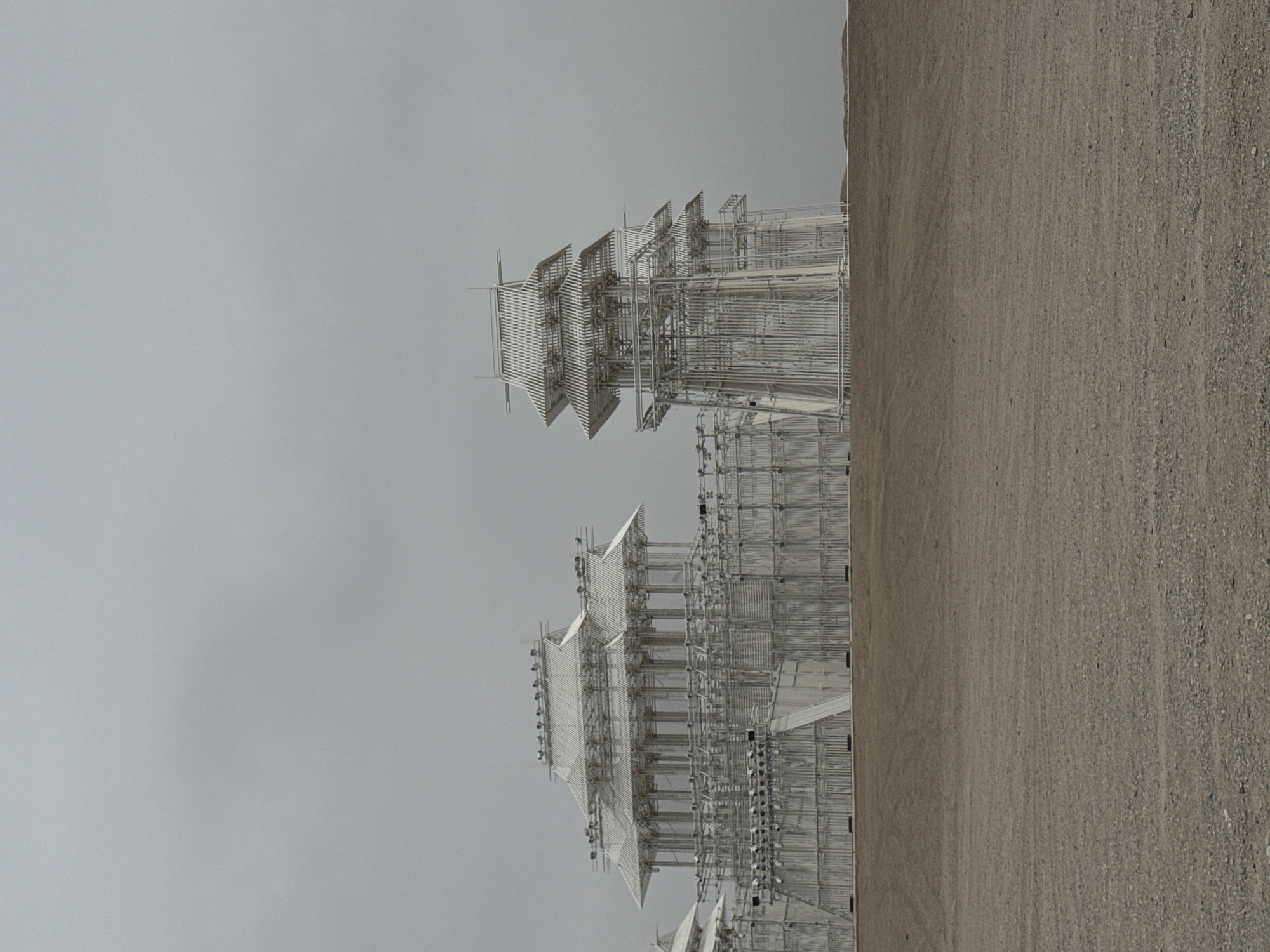 The image shows a large, modern architectural structure composed of multiple tiered and interconnected sections. The structure appears to be constructed of metal and possibly wood, with a dense network of beams and panels. The overall design evokes a sense of abstract geometric form and complexity. The structure is positioned on a flat, gravelly surface under an overcast sky. It is located in Guazhou County, China.