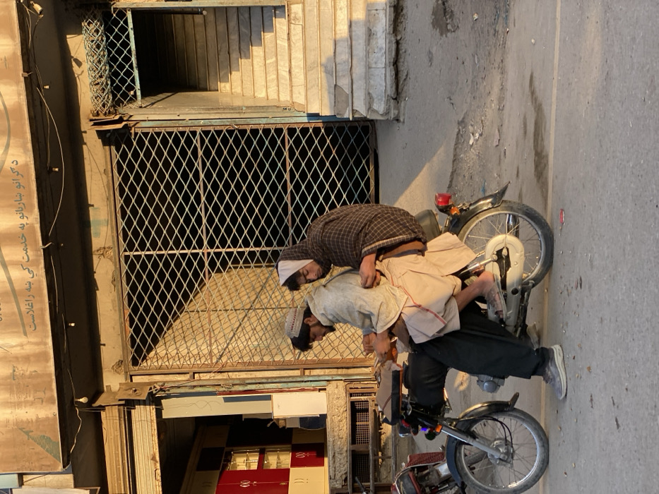 The image depicts a street scene in Kandahar, Afghanistan, during the golden hour, characterized by warm, directional sunlight. Two males are positioned on a stationary, dark-colored motorcycle on an asphalt street. The male in the driver's seat wears a light-colored cap, a grey or beige tunic over dark trousers, and grey sneakers; his left foot rests on the ground. He is holding a light-colored object in his hands. The passenger, seated directly behind the driver, wears a dark plaid shawl covering his head and upper body, over light-colored trousers, and is barefoot, with his right foot resting on a motorcycle footpeg. He appears to be looking down at his hands or at the object held by the driver.
The background consists of two adjacent building facades. To the left, a storefront with a vertical architectural element reveals red and beige storage or display units visible through an opening. To the right, a building features a large, dark, diamond-patterned metal security gate, partially closed. Above this gate, a light-colored sign displays Pashto script, which translates to "Dear guests, welcome to the service," and exposed electrical wires with small bulbs. Adjacent to the gate on the right, an exterior staircase with worn, light-colored steps leads upward. The street surface is uneven, and prominent shadows are cast towards the right side of the frame.