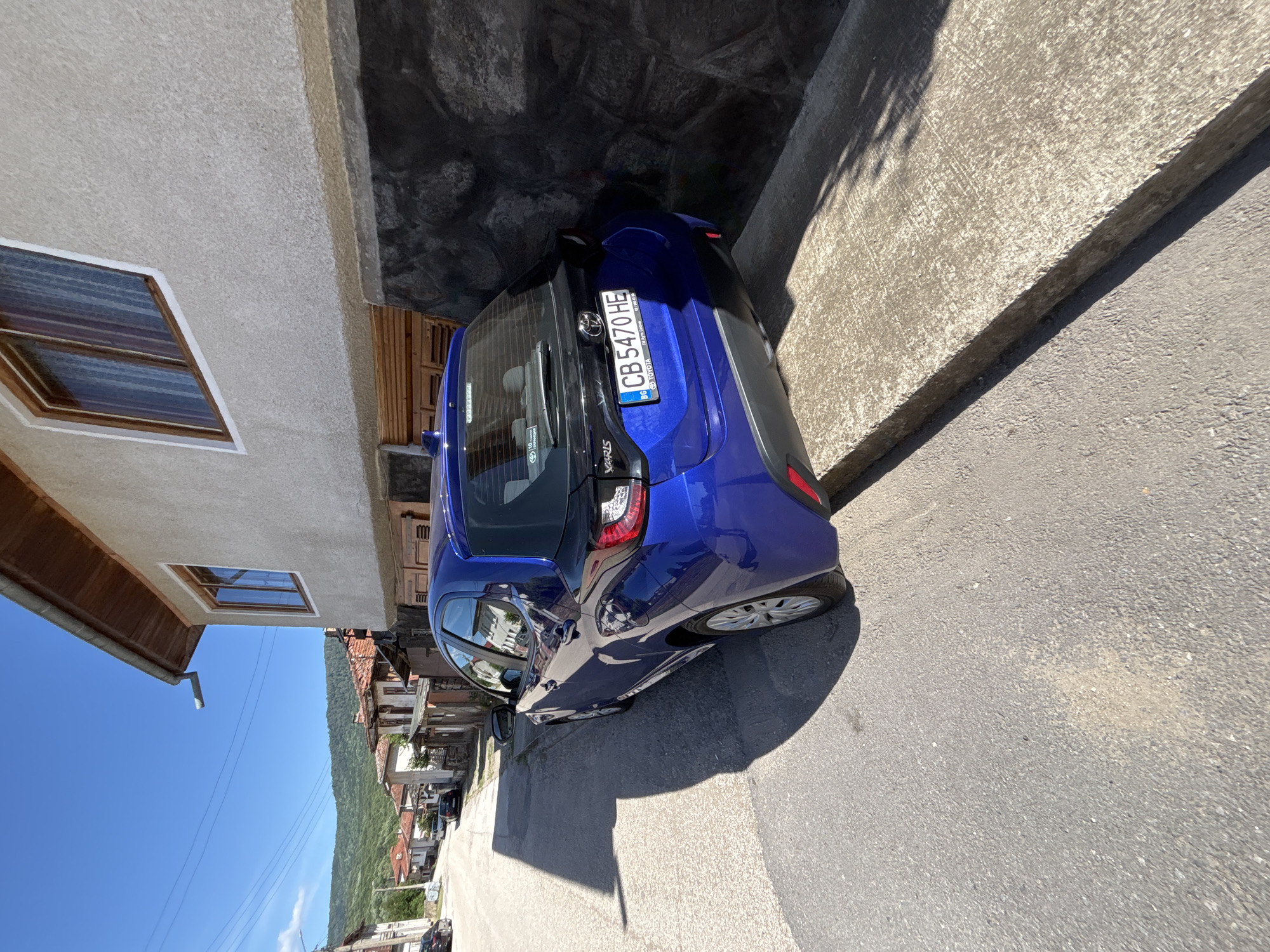 The image shows a blue Toyota Yaris parked on a narrow street in what appears to be Balgarene, Bulgaria. The car is close to two buildings, one with a window and another of stone. The license plate is visible: CB 5470 HE. Beyond the car, the street extends towards a hill and other buildings. The sky is clear and blue.