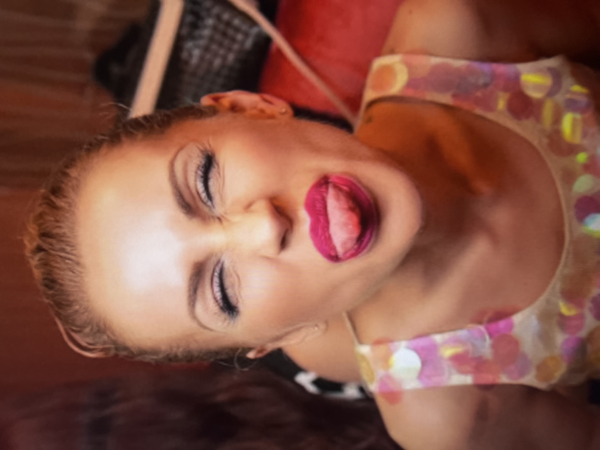 A close-up, medium shot shows a woman with her eyes closed and her tongue sticking out. She is wearing a colorful sequined top. The background is blurry and appears to be indoors, with red and dark tones. No specific location in Qarağandı city administration, Kazakhstan, can be identified from the image.