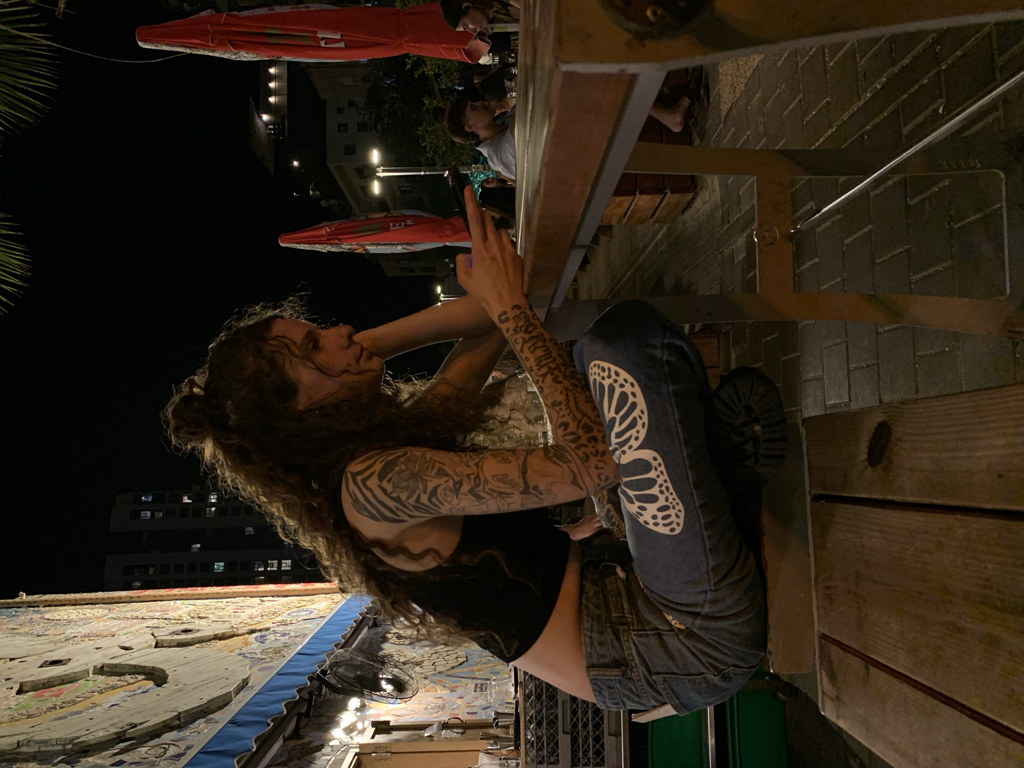 A sideways nighttime shot, possibly taken in Tel-Aviv, Israel, shows a woman with curly brown hair and multiple arm tattoos sitting at a wooden outdoor table. She wears a black top and denim overalls with a butterfly design on one leg. The table is part of an outdoor dining setup with red umbrellas visible in the background. Other people are present at the table. In the foreground, there is a mosaic-covered surface with a fan. Buildings with lit windows are visible in the dark background, indicating an urban setting.