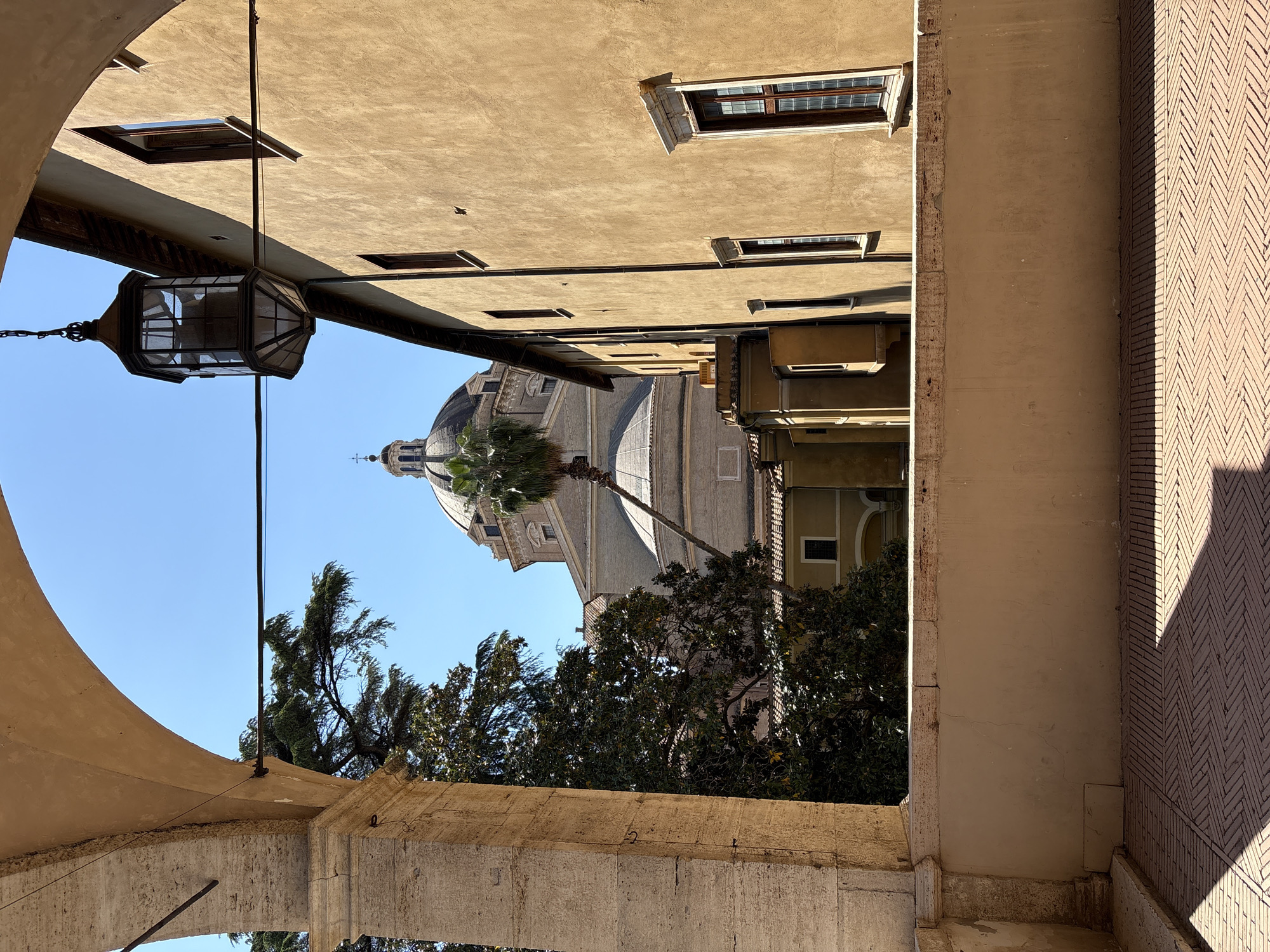 This media file presents a sunlit architectural street scene, viewed from beneath a stone or concrete archway.
**Subjects:** The central subjects include a narrow street or alley flanked by multi-story buildings, a prominent church dome in the background, and various trees. In the immediate foreground, a large, ornate metal lantern with glass panels hangs from the underside of the archway, suspended by a chain and rod. To the left, a tall building with a light-colored, possibly stucco or plaster, facade features several rectangular windows with wooden frames, some slightly ajar. Beyond this, a grand domed structure, characteristic of a church or basilica, rises against the sky, topped with a smaller cupola and a cross. Several tall palm trees and other dense green foliage are visible in front of and around the dome. On the right side of the street, another building facade appears, partially shadowed, with a distinctive herringbone brick pattern visible on a lower wall or ground section.
**Activity or event taking place:** The image captures a static, uninhabited scene. No discernible activity or event is occurring.
**Notable details:** The scene is illuminated by strong, clear daylight under a bright blue sky, indicating fair weather and likely a time between late morning and mid-afternoon, given the cast shadows. The architectural elements, particularly the large dome, the aged building facades, and the presence of palm trees, strongly suggest a Mediterranean urban setting, consistent with Rome, Italy. The detailed design of the hanging lantern adds an element of historical charm. No visible text is present in the image. The overall impression is one of a quiet, historic European street.