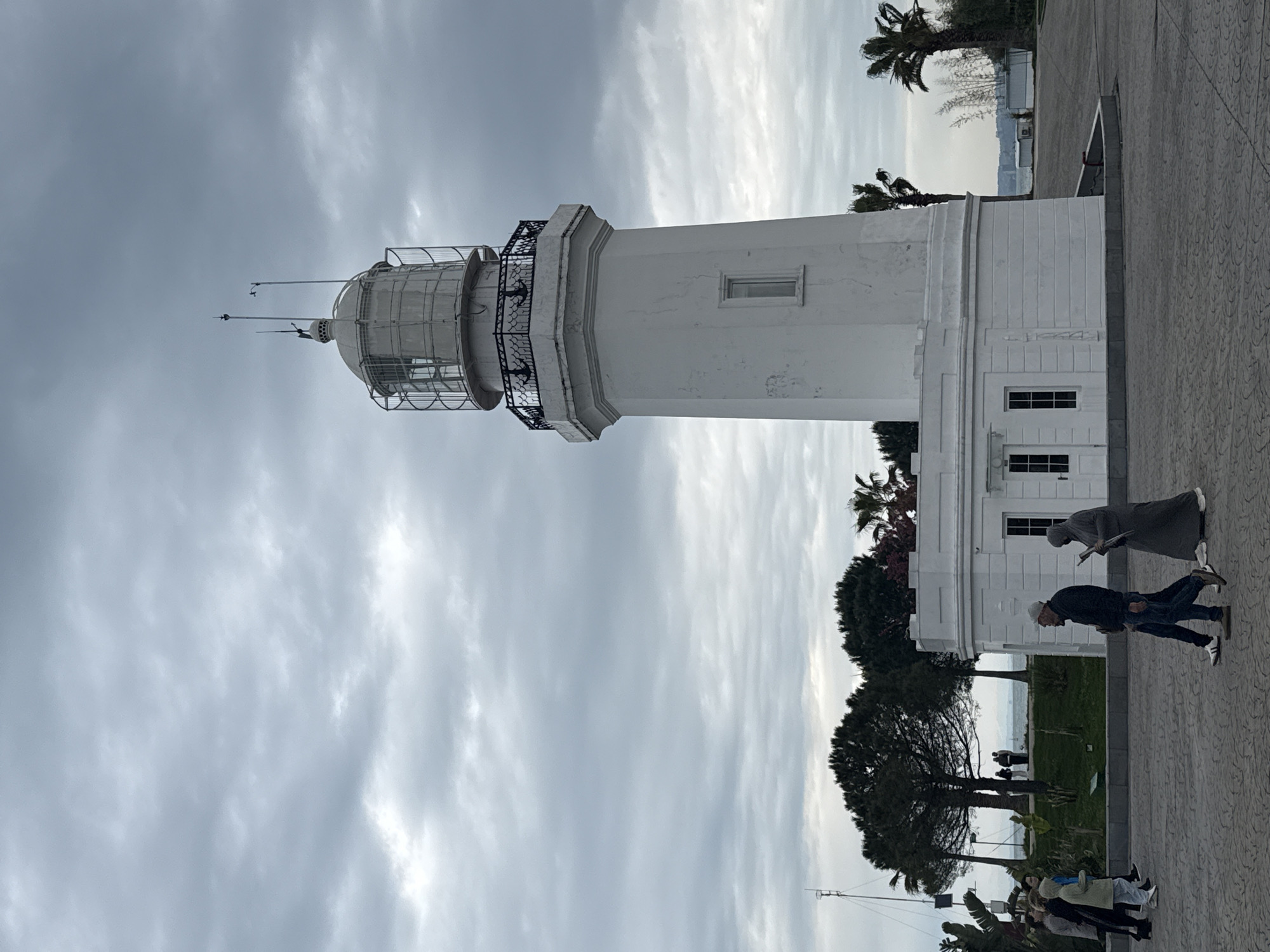 A tall, white lighthouse stands prominently on a cloudy day. Two people are walking in front of the lighthouse. Other people are standing in the foreground. The lighthouse is located in Batumi, Georgia. Trees and palm trees surround the lighthouse. The ground is paved with a patterned stone.