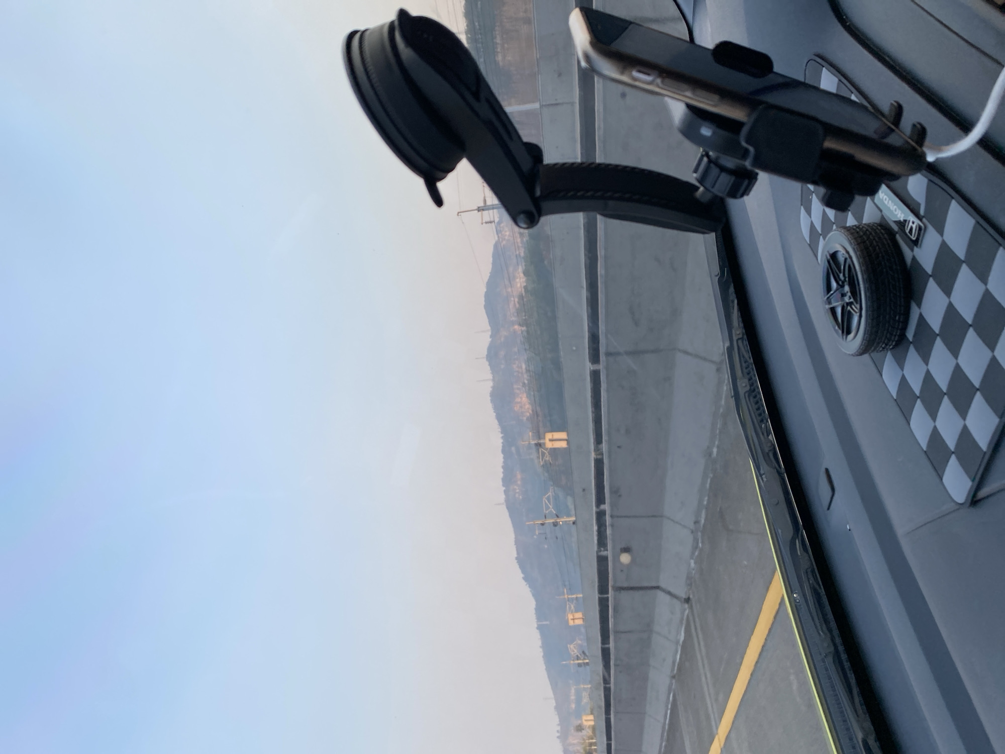 The image shows a view from inside a car, looking out through the windshield. The car is on a highway, with a concrete barrier separating the lanes. In the distance, there are hills with some structures on them, possibly utility poles or antennas. The sky is a clear blue, fading to a lighter shade near the horizon.
Inside the car, on the dashboard, there are several objects. A black phone mount is prominently visible, holding a smartphone. Next to the phone mount is a small, decorative car wheel. Underneath these items is a checkered mat, and part of a car's dashboard is also visible. A yellow line is visible on the road to the left, indicating a lane marking. The overall impression is that of a car traveling on a highway during the day, with a clear view of the landscape ahead. There are no people visible in the image. The location of Nyalindung, Indonesia is not determinable from the visual information provided.