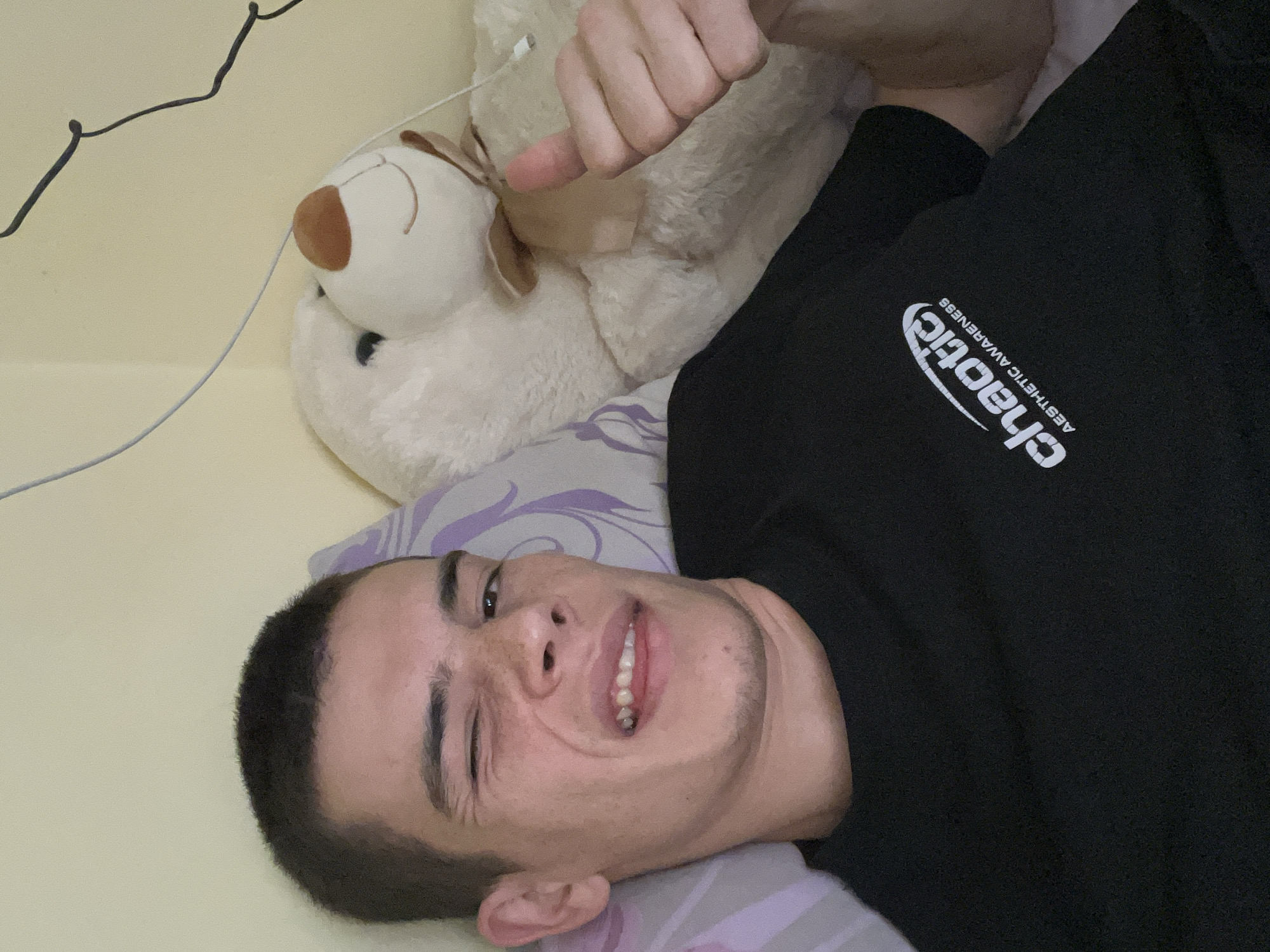 A young adult male is lying on a bed, partially propped on a pillow with a lilac and white swirl design. He is fair-skinned with short, dark hair and a slight stubble. He is wearing a black sweatshirt with the word "chaotic" printed on it. He is looking at the camera with a slight smile and his right eye is squinted. A large, light-colored teddy bear is positioned behind him. A thin, white wire or cable is visible in the upper left corner of the image. The bed has a light yellow sheet. The location is unknown, and there is no information indicating that it is in the city of Plovdiv, Bulgaria.