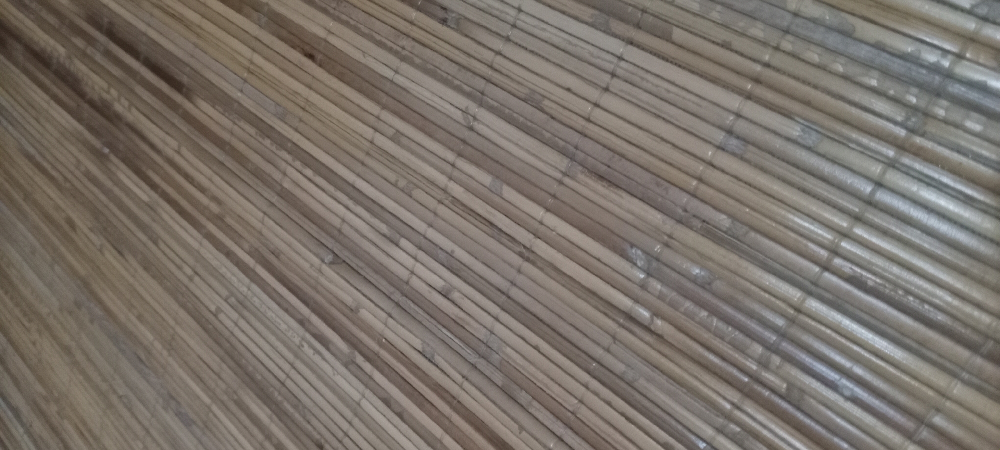 The image displays a close-up, angled view of a woven bamboo surface, likely a floor mat or part of a furniture piece. The bamboo stalks are thin, uniformly cut, and laid parallel to each other, creating a textured pattern. The color of the bamboo ranges from light tan to a medium brown, with some stalks exhibiting a slightly darker hue, suggesting variations in age or material. The surface appears to have a semi-gloss finish, reflecting light in diagonal streaks across the stalks, particularly on the lower half of the image. There are no people, animals, or other objects visible in the frame. The scene is devoid of any clear environmental indicators that would place it within a specific urban or natural setting, and therefore, its location in Sindangwangi, Indonesia, cannot be confirmed from the visual information alone.