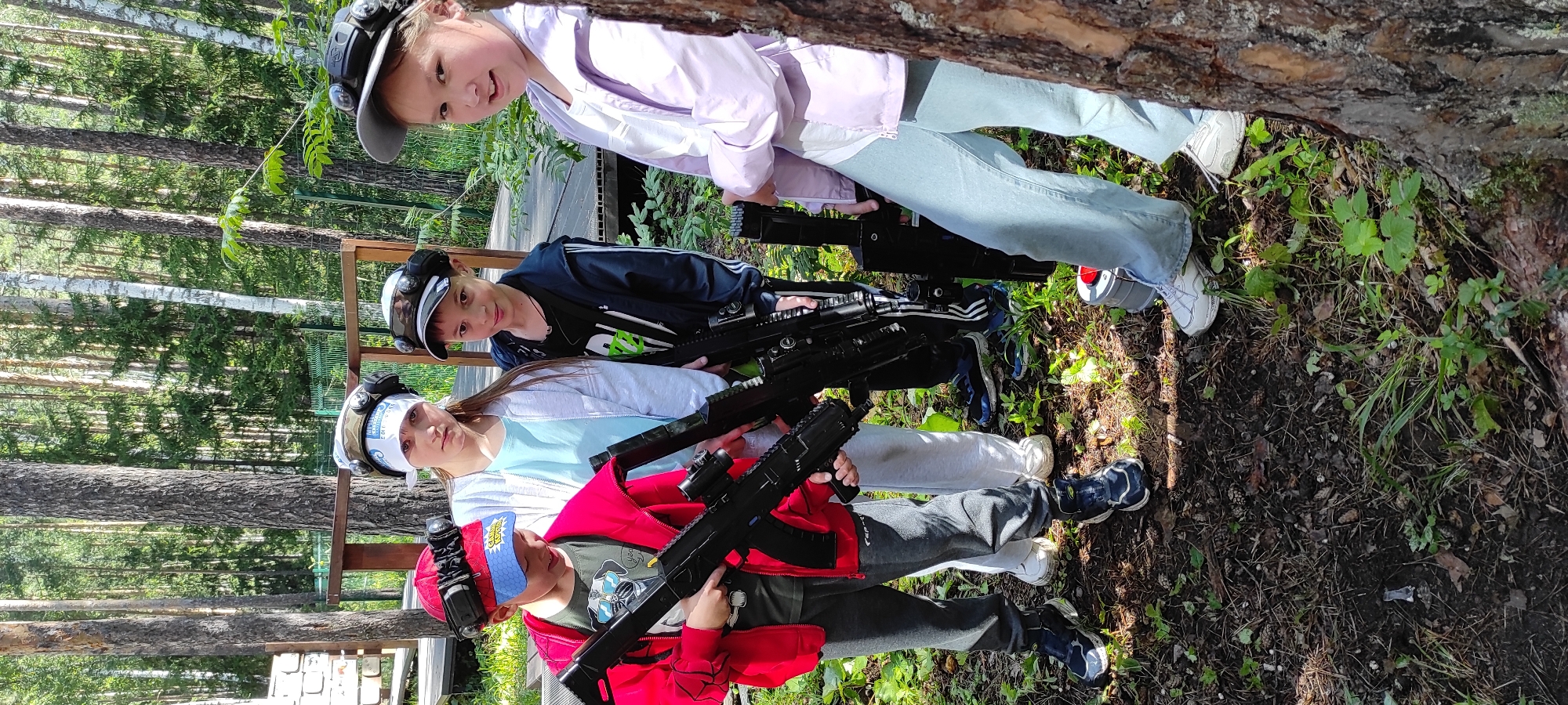 Four children, two boys and two girls, are positioned in a wooded area, likely a laser tag arena. They are wearing laser tag vests and headgear with illuminated sensors. Each child is holding a black laser tag gun. The children are dressed in casual clothing. The environment consists of trees, undergrowth, and a wooden platform in the background. The ground is covered with dirt, pine needles, and some green foliage. The lighting suggests it is daytime. The presence of the laser tag equipment and setup indicates a recreational activity.