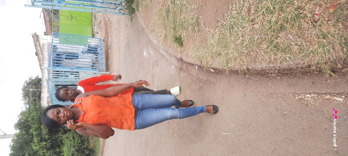 Two women are walking on a paved path next to a grassy area. The woman in the foreground is wearing an orange top and blue jeans and looking down while touching her face. Behind her, another woman is wearing a red long-sleeved shirt. They are walking in front of a building with a blue metal gate. The background includes some trees and a building. "Police kombo" is written at the bottom right corner.