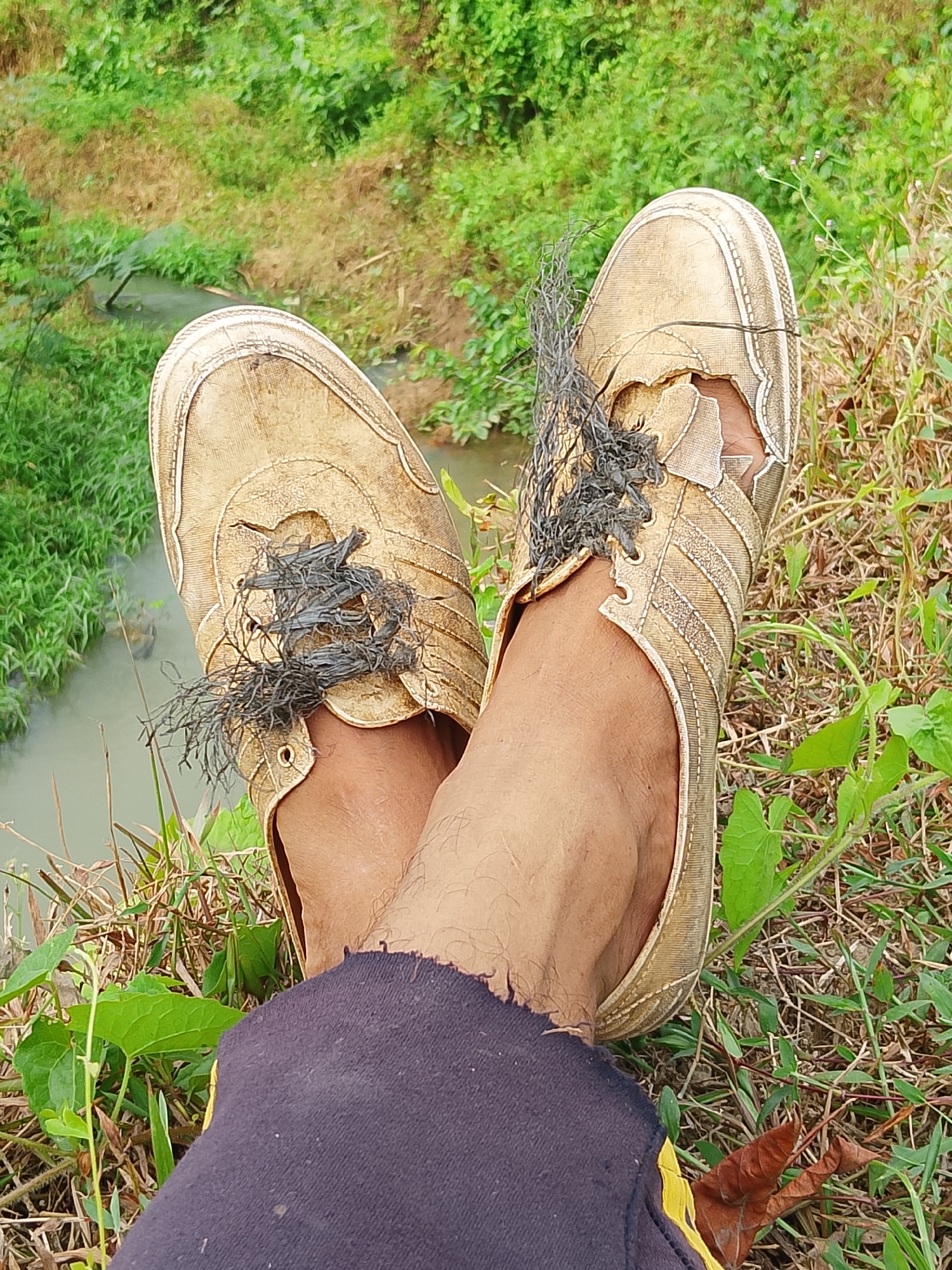 A close-up, overhead shot shows a person's legs and feet in worn-out gold sneakers, resting on grass near a small, muddy stream. The sneakers are ripped, with frayed laces spilling out. The legs are clad in dark pants with ripped hems. The background is a lush green, overgrown area with trees and foliage. The location appears to be an outdoor, undeveloped area, possibly in a rural part of Indonesia, given the vegetation and stream.