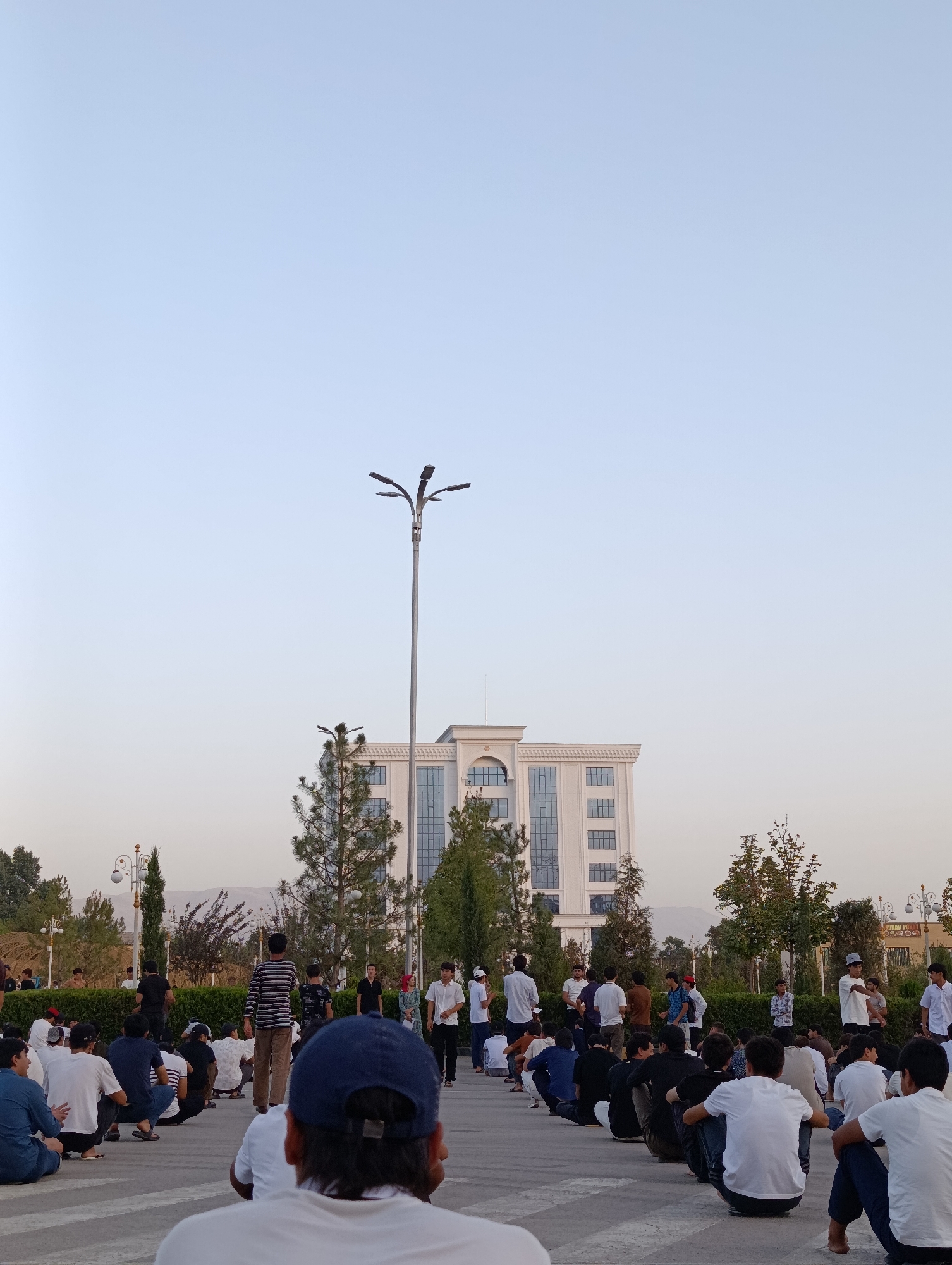In Danghara, Tajikistan, a large group of men are gathered in a paved outdoor area in front of a modern, multi-story building with a light-colored facade and numerous windows. The men are mostly sitting on the ground in rows, facing towards a large streetlamp with multiple lights. Several trees are scattered throughout the scene, with some taller coniferous trees near the building and shorter, bush-like hedges along a walkway. The sky is clear and pale blue, indicating it is likely late afternoon or early evening. The overall atmosphere appears calm, with people casually interacting or observing.