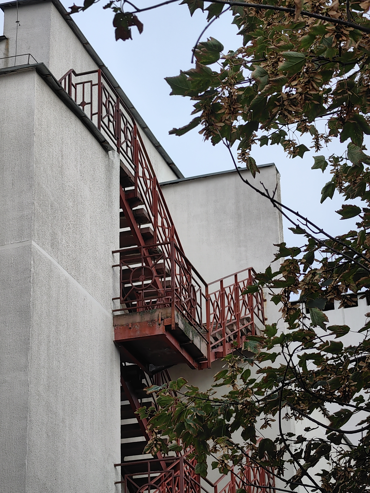 An exterior shot of a white apartment building with a red metal fire escape. The fire escape is attached to the side of the building and extends downwards. It is made of metal railings and has several landings. The building appears to be in an urban setting, with trees and foliage visible in the foreground. The sky is overcast. The location is Minsk, Belarus.