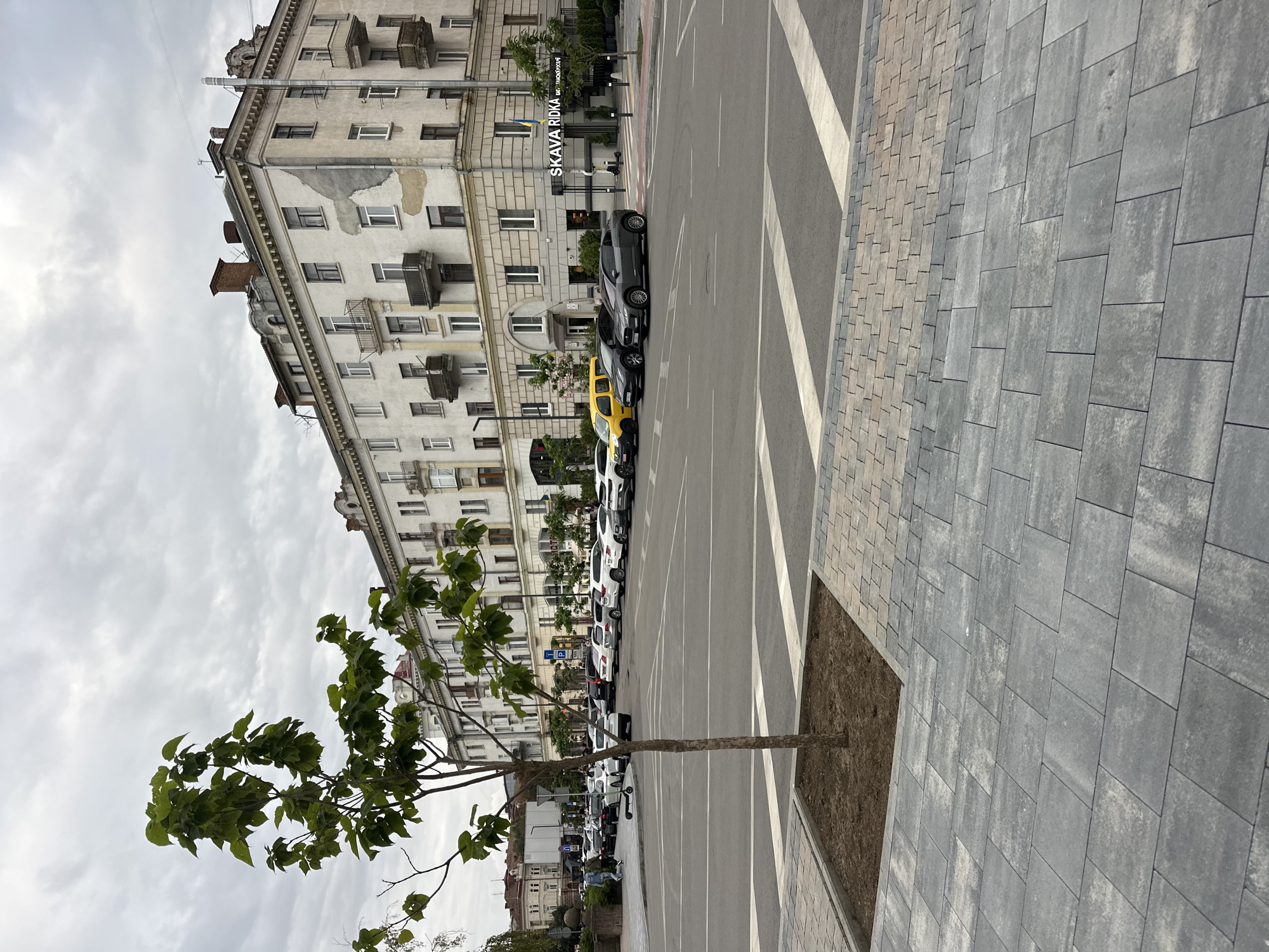 The image depicts a street scene in the city of Ternopil, Ukraine, specifically at the intersection of Shevchenko Street and the Ternopil Pond embankment. A young tree stands in the foreground, planted in a square garden bed. The street itself is paved with grey interlocking stones, transitioning to asphalt further down the road. A crosswalk is visible across the asphalt.
In the background, a large, multi-story building with a cream-colored facade and numerous windows stands prominently. The building exhibits signs of age, with some peeling paint and weathered balconies. A sign on the building reads "SKAVA RIDKA" in large letters, indicating a business located there. Several cars are parked along the street, including a yellow hatchback, a dark sedan, and a grey coupe. A black electric scooter is parked near the building.
The sky is overcast with grey clouds, suggesting a cloudy day. The overall impression is that of an urban environment with a mix of old architecture and modern elements.