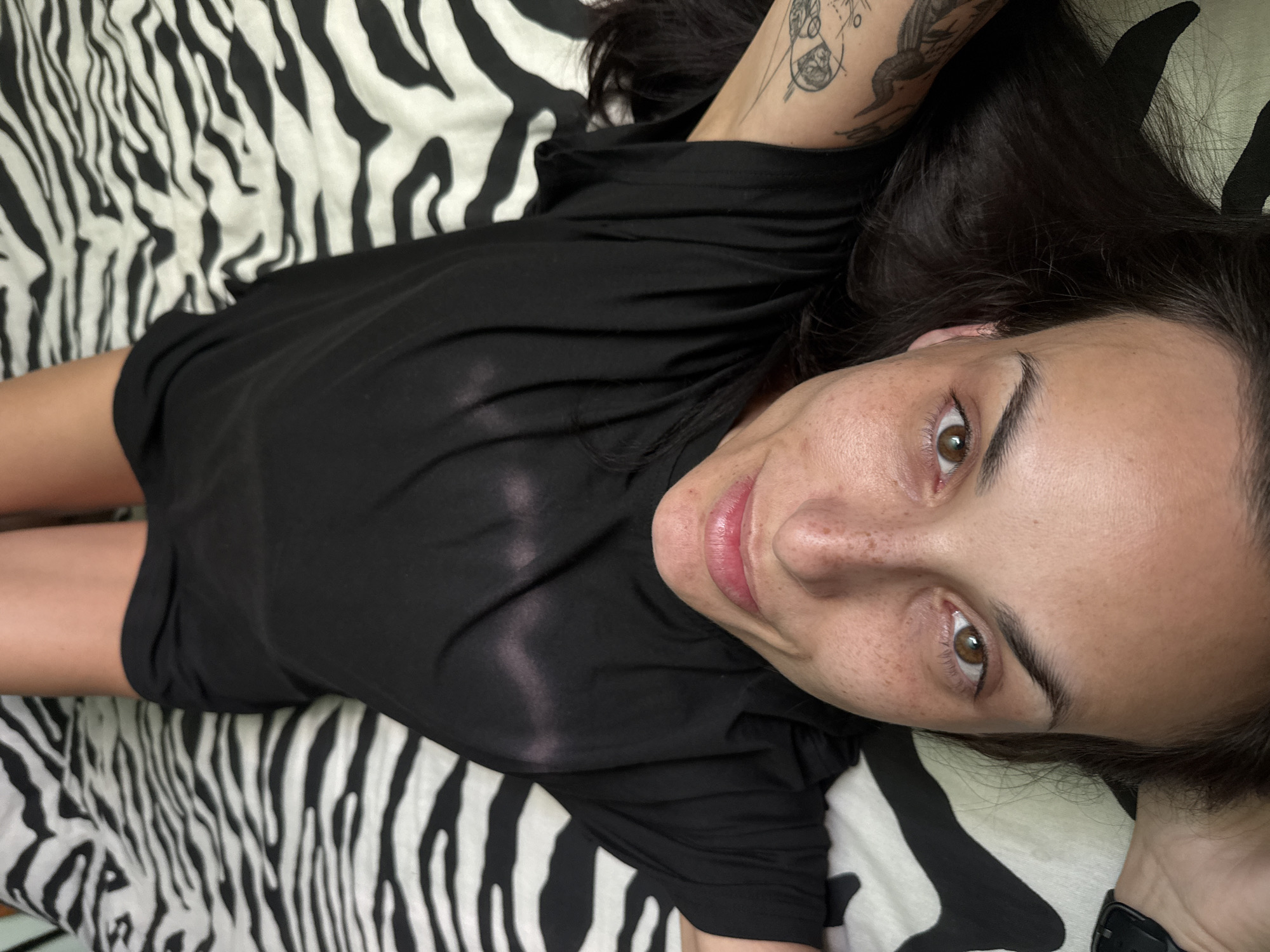 A fair-skinned woman with brown eyes and brown hair lies on a black and white zebra-patterned bedspread. She wears a black t-shirt and short black shorts. Her left arm is visible and has a tattoo on the forearm. Her right arm is partially visible. Her expression is a slight smile. The photograph's location cannot be determined from the provided image.