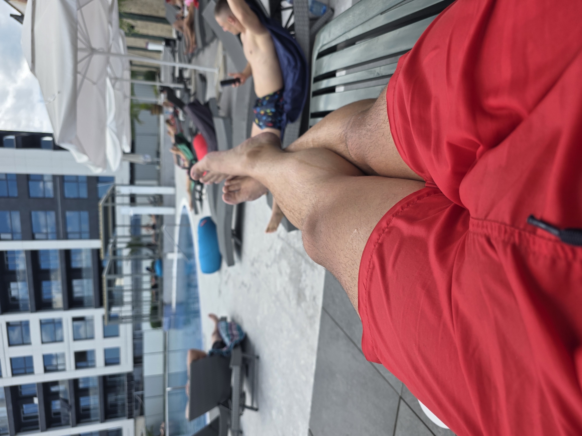 The image depicts a rooftop pool area at a modern apartment building, possibly in Sofia, Bulgaria. In the foreground, a person's legs, clad in red swim trunks, are crossed, suggesting a relaxed posture on a lounge chair. In the mid-ground, other individuals are seen lounging on chairs by the pool, some reading or looking at their phones. The pool itself is visible, with the water reflecting the sky and surrounding structures. The background is dominated by the facade of a multi-story apartment building, featuring a grid of windows and balconies. White umbrellas are set up to provide shade around the pool area. The overall scene conveys a sense of leisure and urban living.