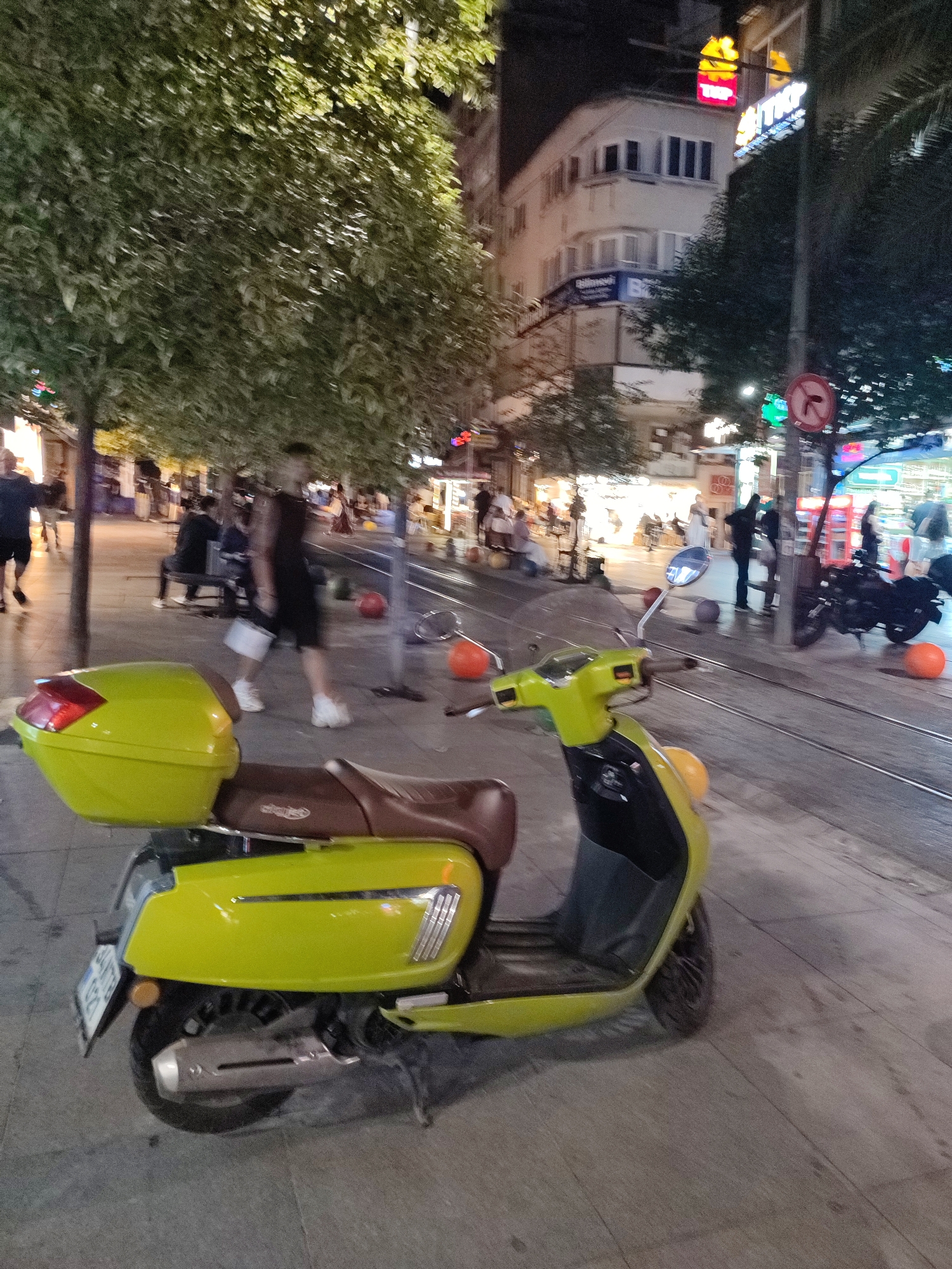 The image depicts a street scene in Kadıköy, Turkey, at night. A bright yellow scooter is parked in the foreground, facing left, with its rear storage compartment visible. The scooter has a brown seat and a license plate. In the background, several people are walking along a sidewalk lined with trees. Some individuals are seated on benches, and others are standing. Buildings with illuminated windows line the street, suggesting commercial establishments. Tram tracks are visible on the ground, indicating the presence of a tram line. Several orange spheres are scattered on the ground. The overall atmosphere is one of a moderately busy urban evening.