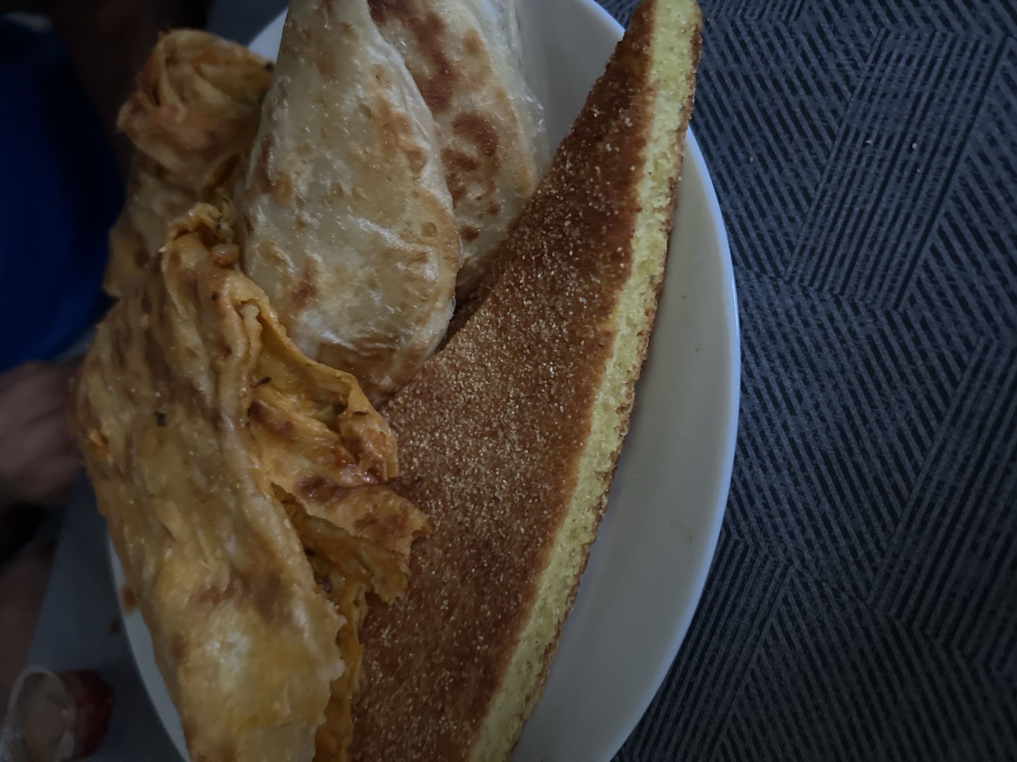 A close-up photograph captures a plate of traditional Moroccan breakfast or snack items resting on a dark grey, patterned table surface in Casablanca, Morocco. The central focus is a large, triangular slice of what appears to be Harsha (semolina bread), displaying a textured, golden-brown crust liberally dusted with semolina, and a dense, pale yellow interior. Behind the Harsha, several pieces of Msemen (flaky, square Moroccan flatbread) are arranged on the white, possibly oval plate. Some Msemen pieces are folded, exhibiting multiple layers and a golden, slightly oily sheen, while one piece appears to be filled with a savory mixture, visible as fragmented contents. Further back on the plate, another style of flatbread, likely plain Rghayef or Msemen, is visible, round and somewhat thinner. In the upper left background, a small, clear plastic ramekin containing a reddish-brown sauce or condiment is partially visible. Blurred human hands, one with a blue sleeve, are present in the background, suggesting individuals are dining or preparing to dine. The lighting is consistent and even.