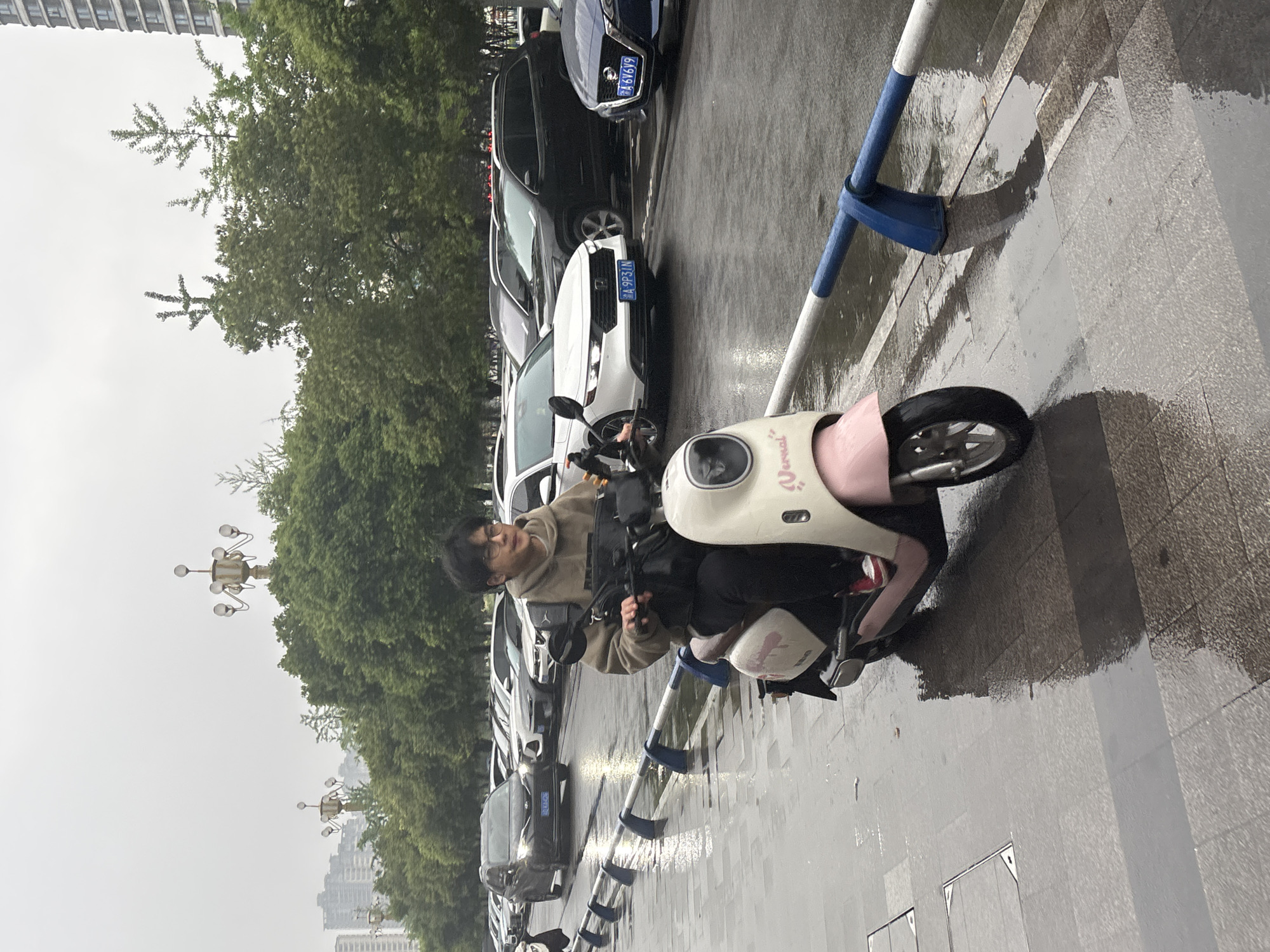 A young man with dark hair and glasses, wearing a tan hoodie, is riding a small, light-colored electric scooter on a wet city street in Jiangjin, China. He is positioned to the right of a blue and white pedestrian barrier. Cars are parked along the left side of the street, and there are trees and streetlights in the background. The sky is overcast. The road is wet and reflecting light.